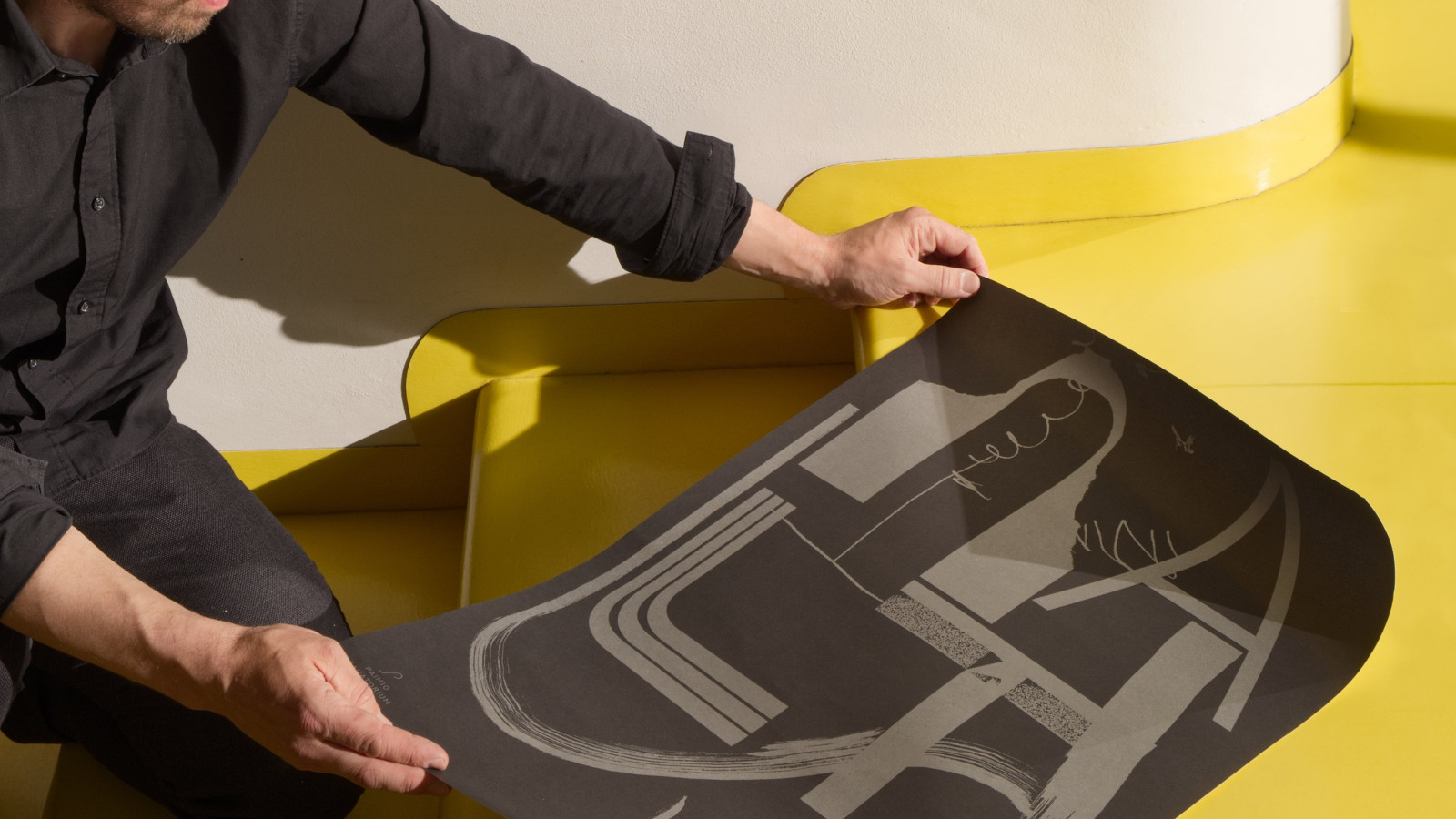 Kuvassa on taiteilijan käsissä tumma juliste, jossa on koukero kuvioita. Takana näkyy keltaista lattiaa.