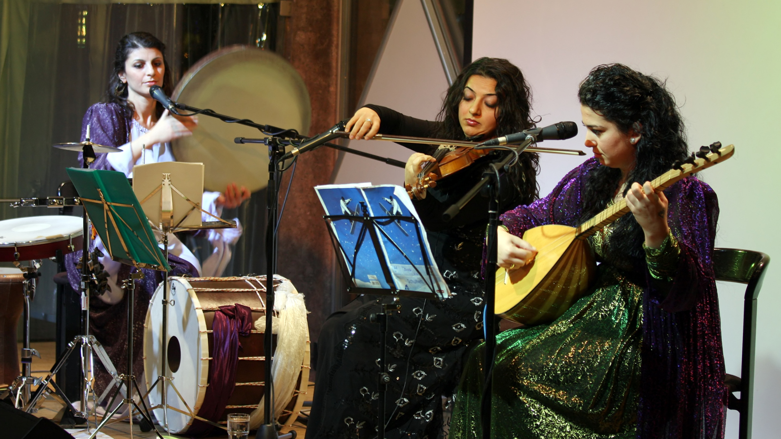 Kuvassa ovat Gûlistan Ensemblen kolme soittajaa esiintymässä ja esiintymisasut ovat tumman kiiltäviä.  Soittimina ovat rummut, viulu ja kastanja.