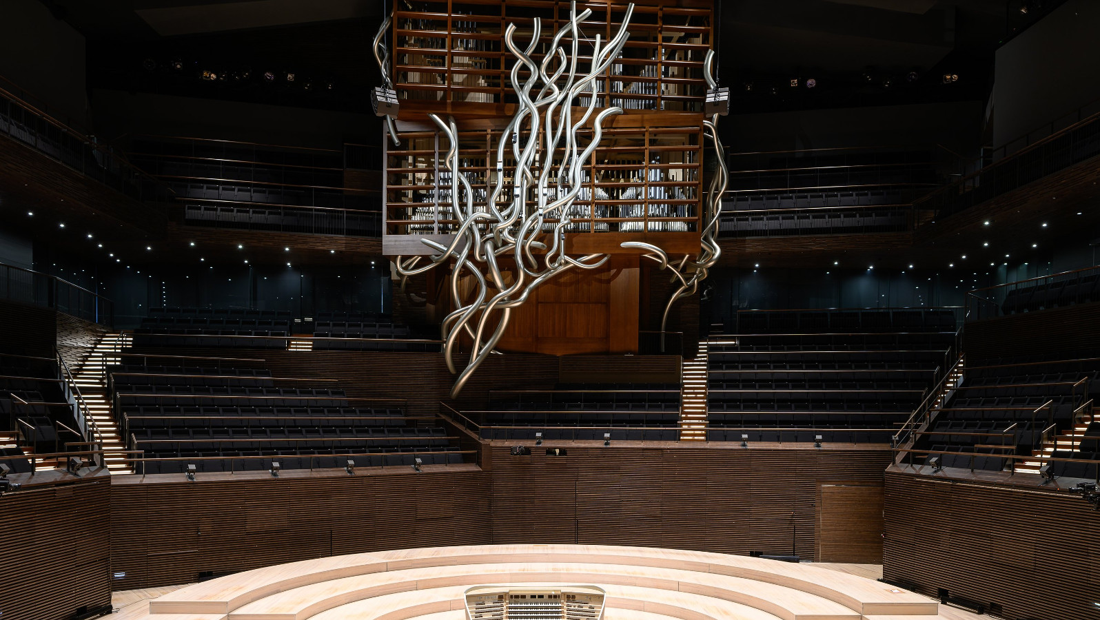 Kuvassa on Musiikkitalon konserttisali tyhjänä ja keskellä ylhäällä on teräksinen kiehkurainen iso koriste. Alla on puinen orkesterialue.  Takana on tumma katsomo ja keskellä kuva ruskeistä uruista.