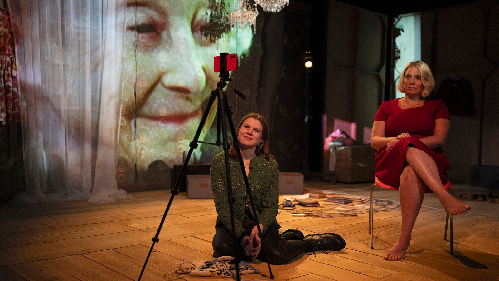 Kuvassa ovat Inke Koskinen lattialla ja Emmi Kaislakari istumassa tuolissa. Edessä on kamera ja taustalla on heijastettuna Seela Sellan kasvot.