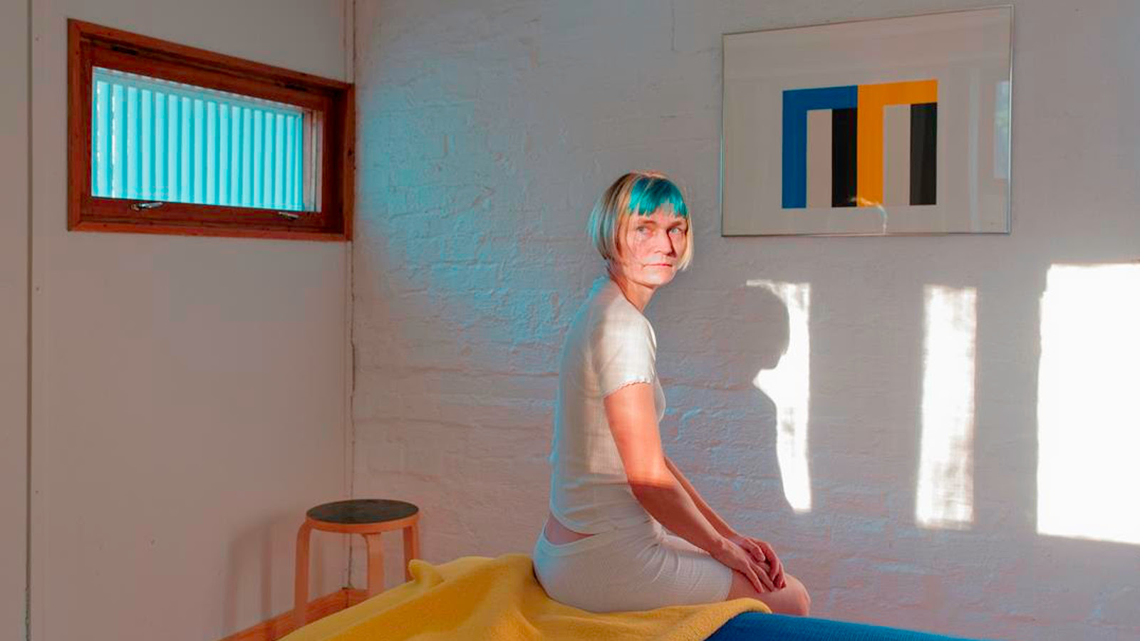 Kuvassa on nainen istumassa Alvar Aallon makuuhuoneessa sängyn reunalla ja sängyssä on keltainen peitto.  Huoneeseen heijastuu valoa sängynpäätyseinän ylhäällä olevasta ikkunasta. Toisella seinälä on taulu, jossa on sinistä, keltaist aja mustaa m-kirjaimen muotoisessa kulmikkaassa kuviossa.