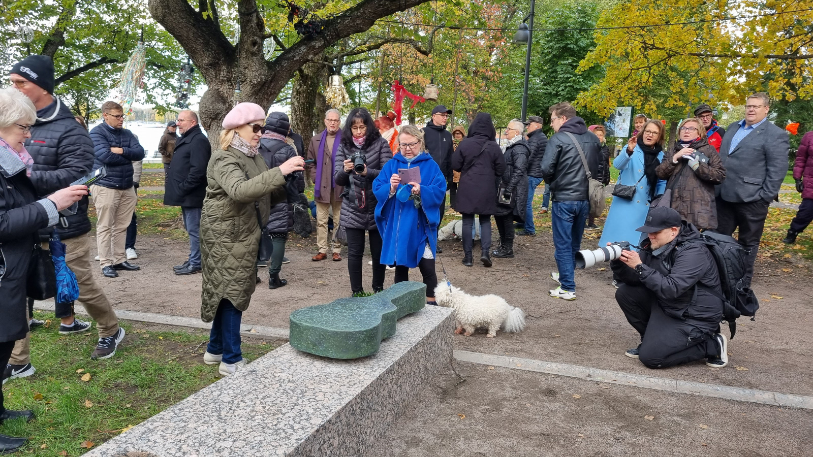 Kuvassa on ihmisiä Lavalla -veistoksen ympärillä ja etualalla on vasemmalla nainen ottamassa kuvaa ja keskellä nainen koiran kanssa sekä oikealla kyykyssä kuvaaja järjestelmäkameran kanssa.   