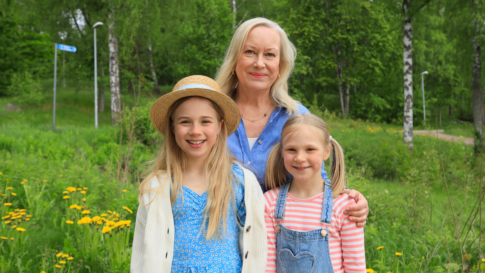 Kuvassa ovat Tiina Nopola ja hänen edessään ovat Eedla Höglund vaalean sinisessä mekossa ja Klaara Höglund vaalean sinisessä haalarissa. He seisovat vihreässä kesäympäristössä.