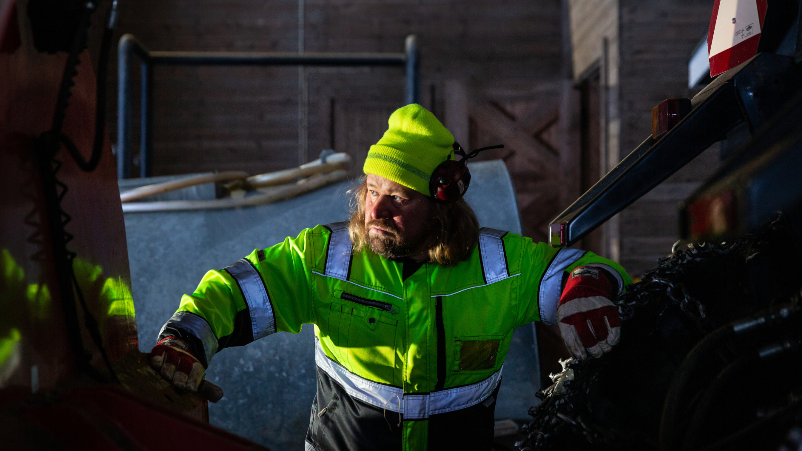 Kuvassa on Janne Kinnunen kirkkaan vaalen vihreä turvahaalari ja pipo päässään. Ympärillä on kkoneita ja latoa.