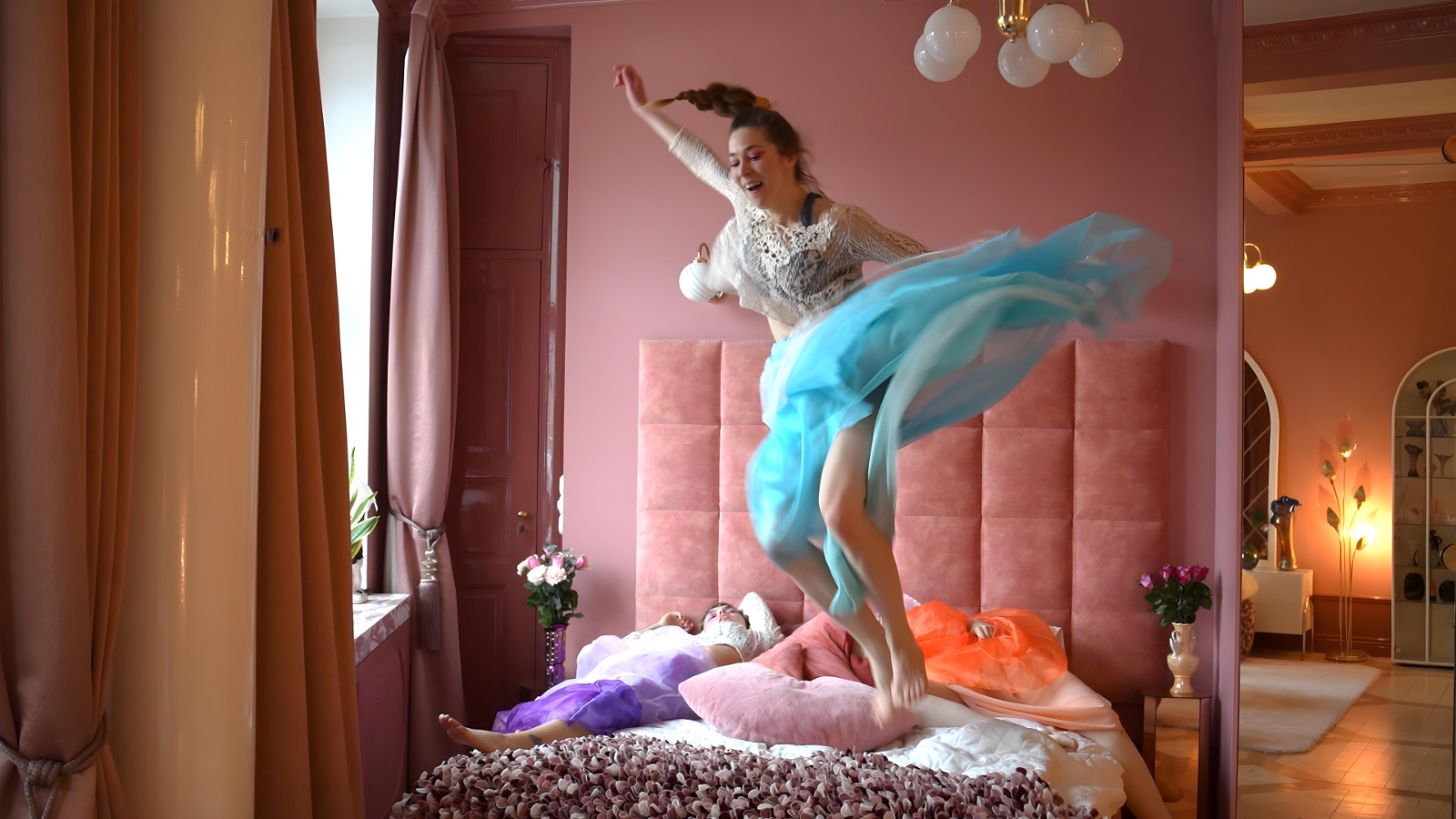 Kuvassa on  on tanssija hyppäämässä sängyn päällä roosan värisessä huoneessa ja taustalla näkyy toista huonetta. Tanssijalla on vaalean sininen tyllihame ja vaalea pusero.