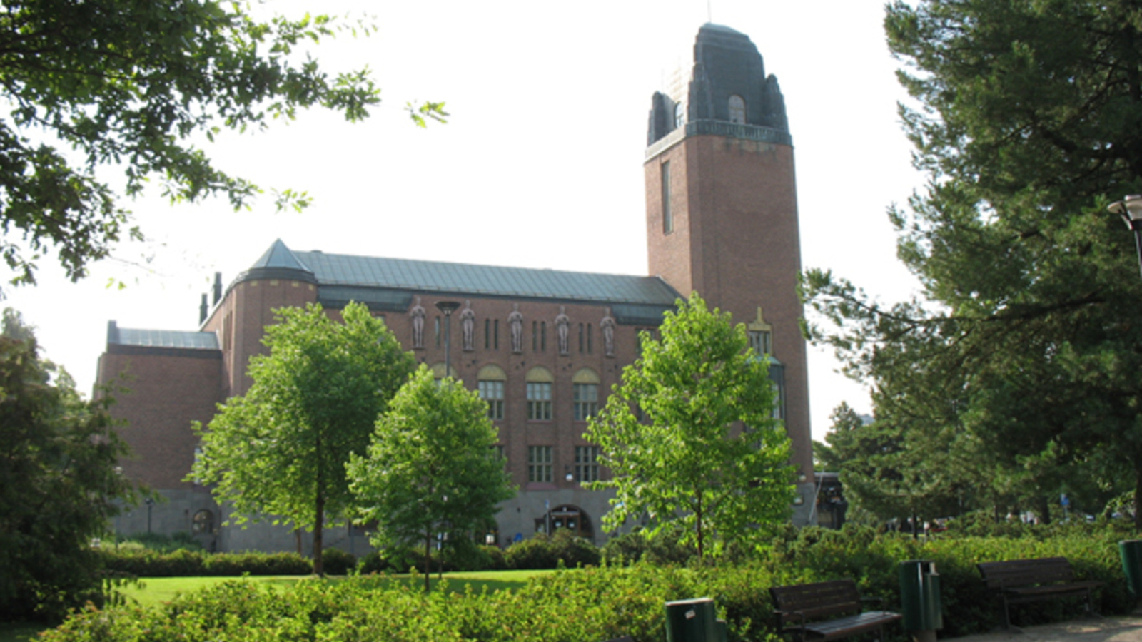 Kuvassa on Joensuun kaupungintalo ja se on punatiilinen kookas rakennus. Sen edustalla on vihreitä. lehtipuita ja pieni puisto.