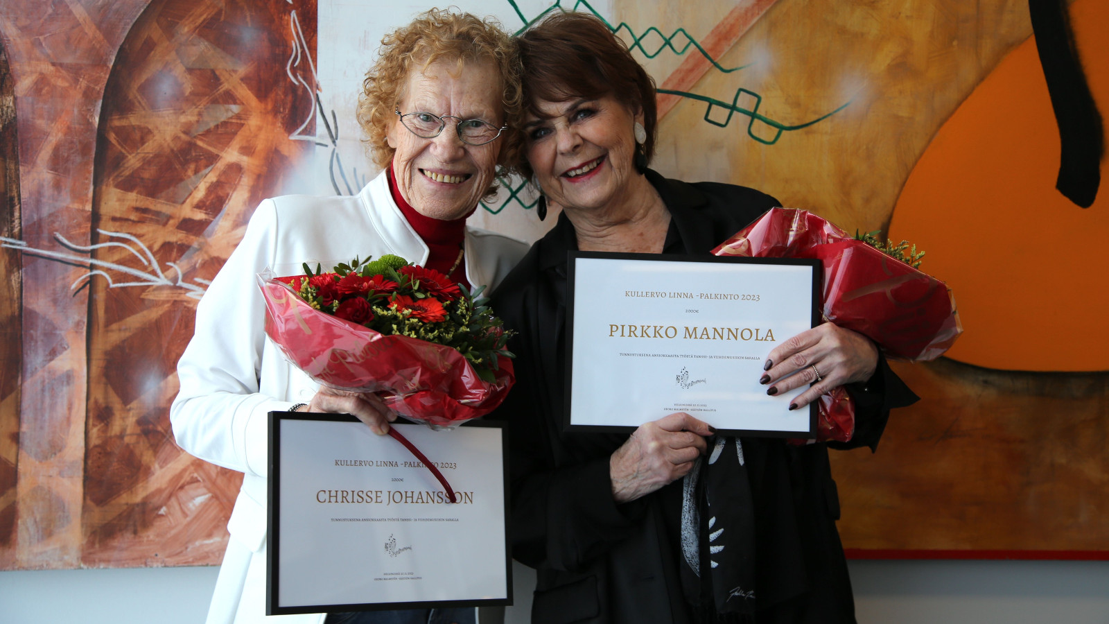 Kuvassa ovat vasemmalla Chrisse Johansson ja oikealla Pirkko Mannola palkintokunniakirjataulut ja kukat kädessään. Johanssonilla on valkoinen jakku ja Mannolalla tumman lila jakku päällään. 