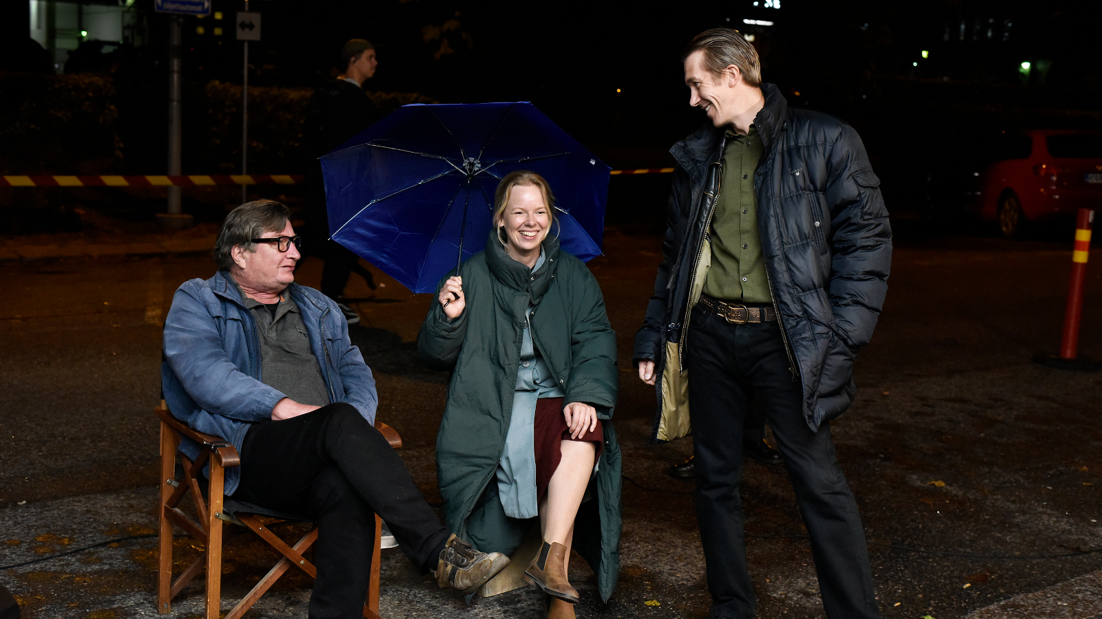 Kuvassa ovat pimeässä istumassa tuoleilla Aki Kaurismäki, Alma Pöysti ja seisomassa Jussi Vatanen. He juttelevat.