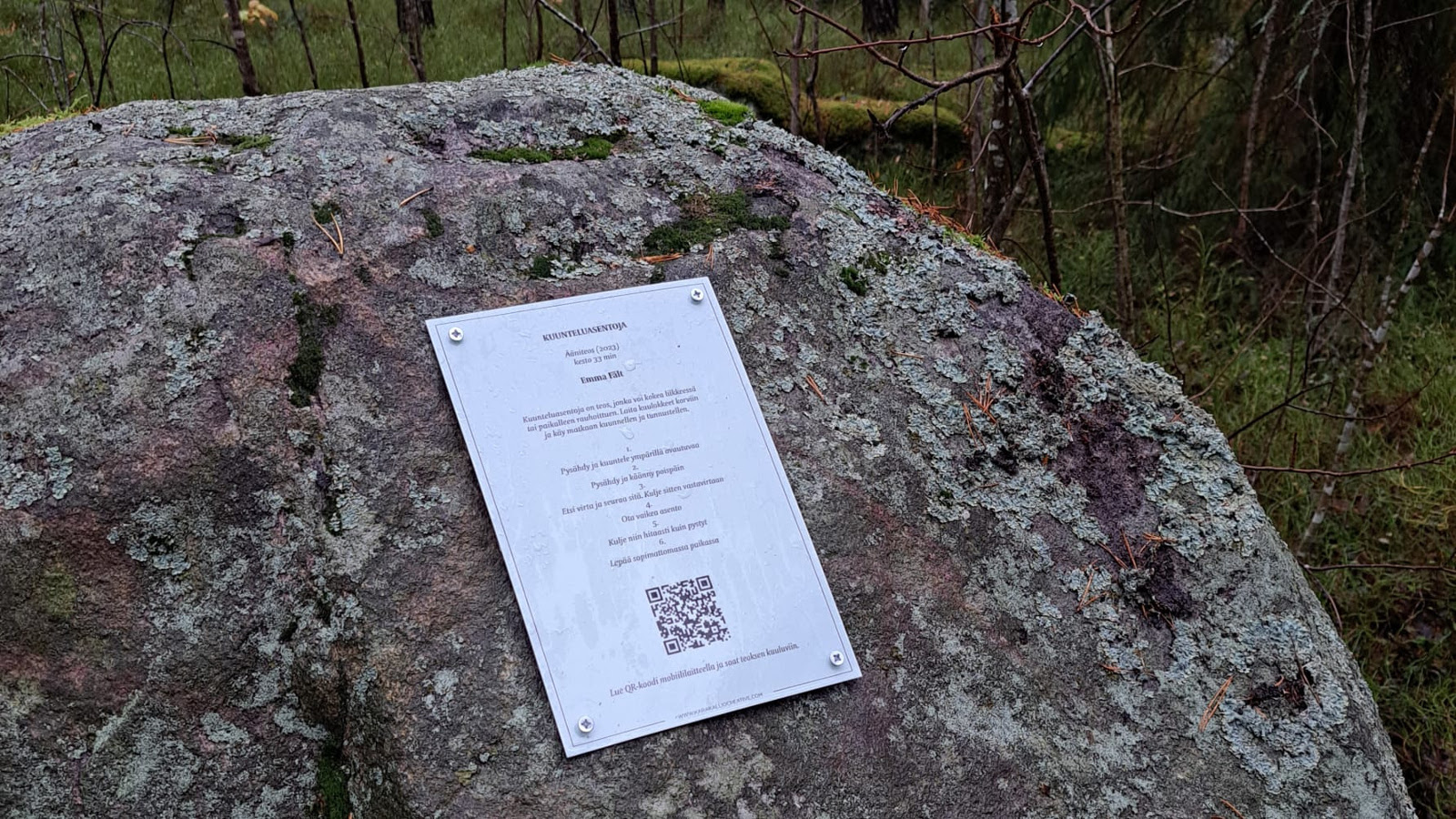 Kuvassa on iso kivi metsässä  ja siihen on kiinnitetty A4-kokoinen, metallinen laatta. Siinä on otsikko Kuunteluasentoja ja ohjetekstiä muutamalla rivillä ja QR-koodi.