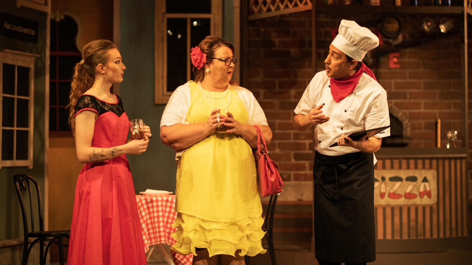 Kuvassa on kaksi naista seisoo keskustelemassa pizzakokin kanssa. Vasemman puoleisella naisella on punainen mekko ja hänen vieressään olevalla keltainen mekko päällään.  Tila on pizzaravintola.