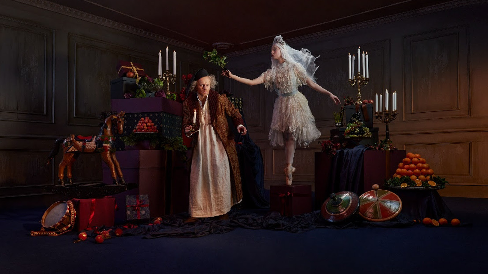 Kuvassa ovat tummassa huoneessa saituri Scrooge ja hänestä vinosti yläpuolella ballerina valkoisessa puvussa. Huoneessa on hedelmiä ja kynttilöitä.