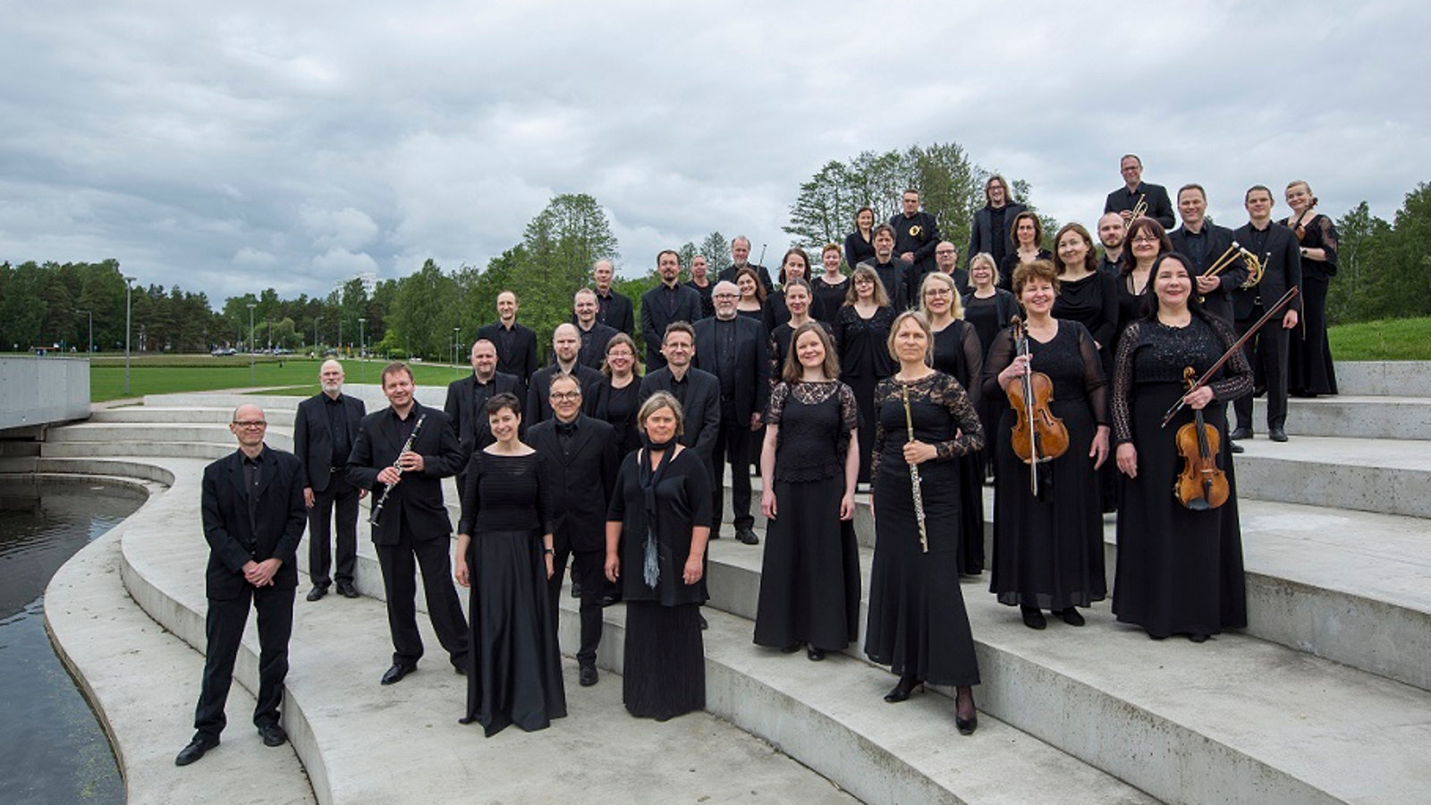 Kuvassa on Tapiola Sinfonietta seisomassa leveillä portailla ja viulisteilla on mustat vaatteet ja soittimet kädessään.