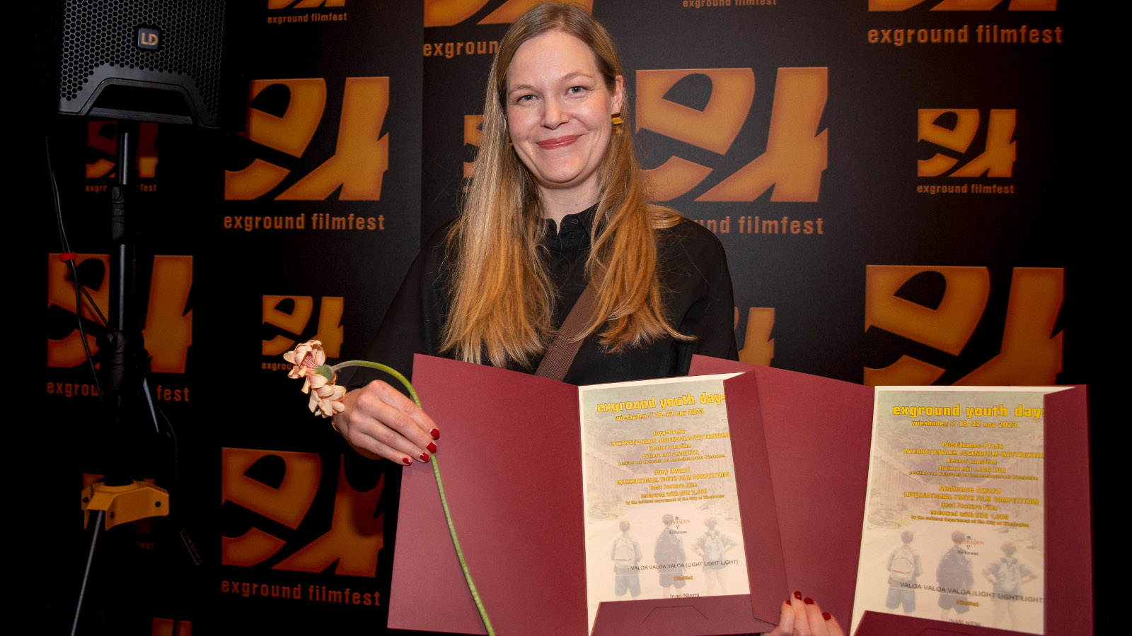 Juuli Niemi seisoo kuvassa keskellä ja hänellä on kahdessa kansiossa kaksi kunniakirjaa. Kansiot ovat burgundin punaisia. Taustalla on filmifestivaalin kelta-mustia julisteita.