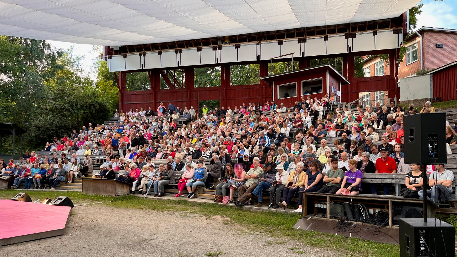 Kuvassa on kesäteatterin katsomo, joka on katettu vaalealla kankaalla. Taustalla on vihreitä lehtipuita. Katsomo on täynnä katsojia.