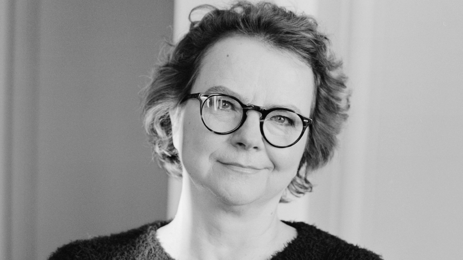 Kuvassa on Laura Kolbe puolivartalokuvassa.  Hänellä on lyhyehköt kiharat hiukset ja silmälasit. Hänellä on päällään tumma villapaita. Kuva on musta-valkoinen. 