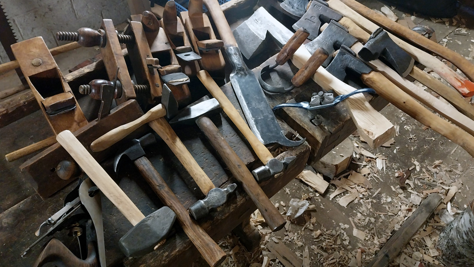 Kuvassa on runsaasti erilaisia työkaluja kuten kirveitä, vasaroita ja lekoja pöydän päällä.