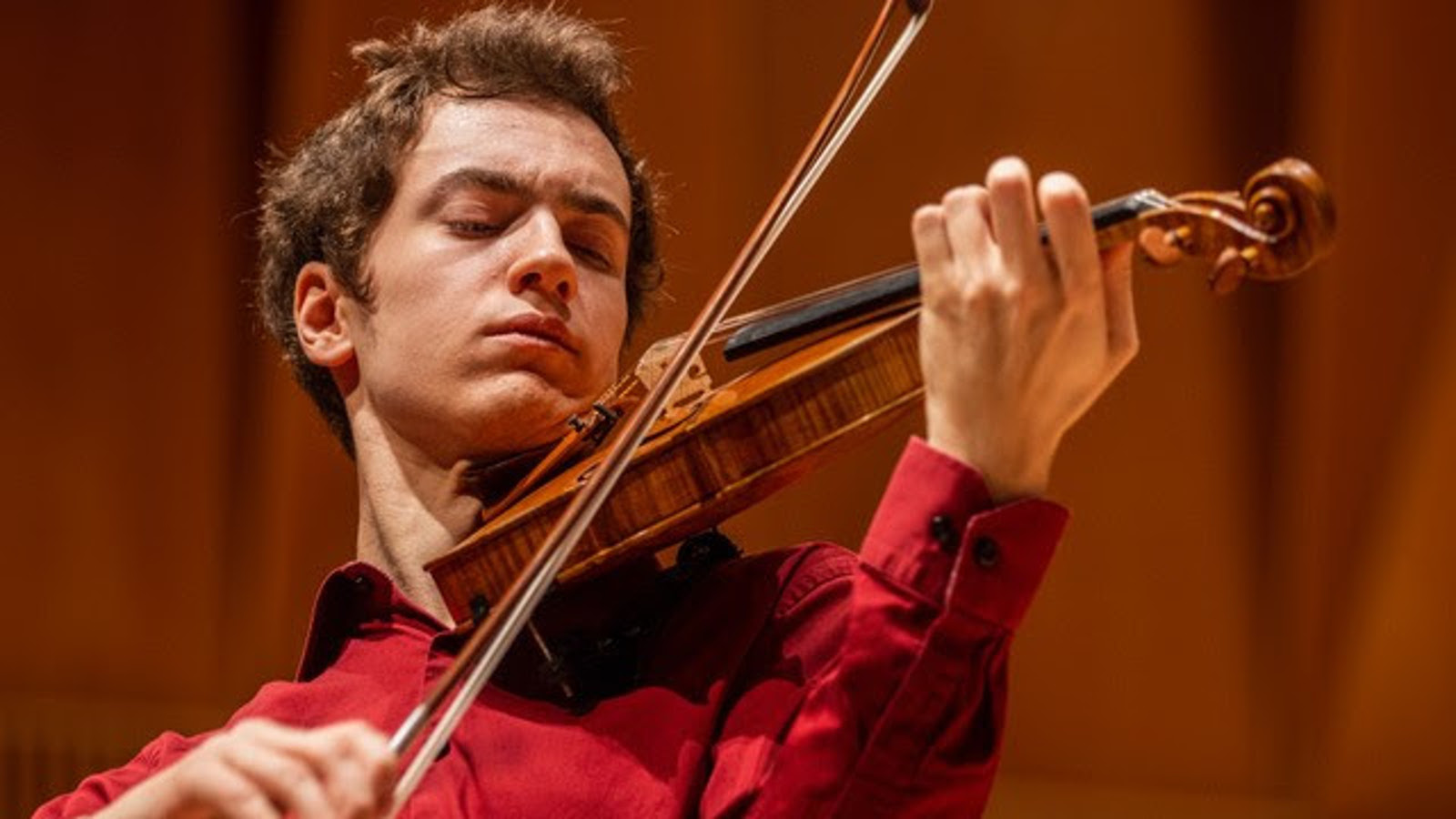 Kuvassa on Adrian Ibañez-Resja soittamassa viulua ja hänellä on punainen paita päällään.  Kuva on puolivartalokuva. Tausta on ruskea.
