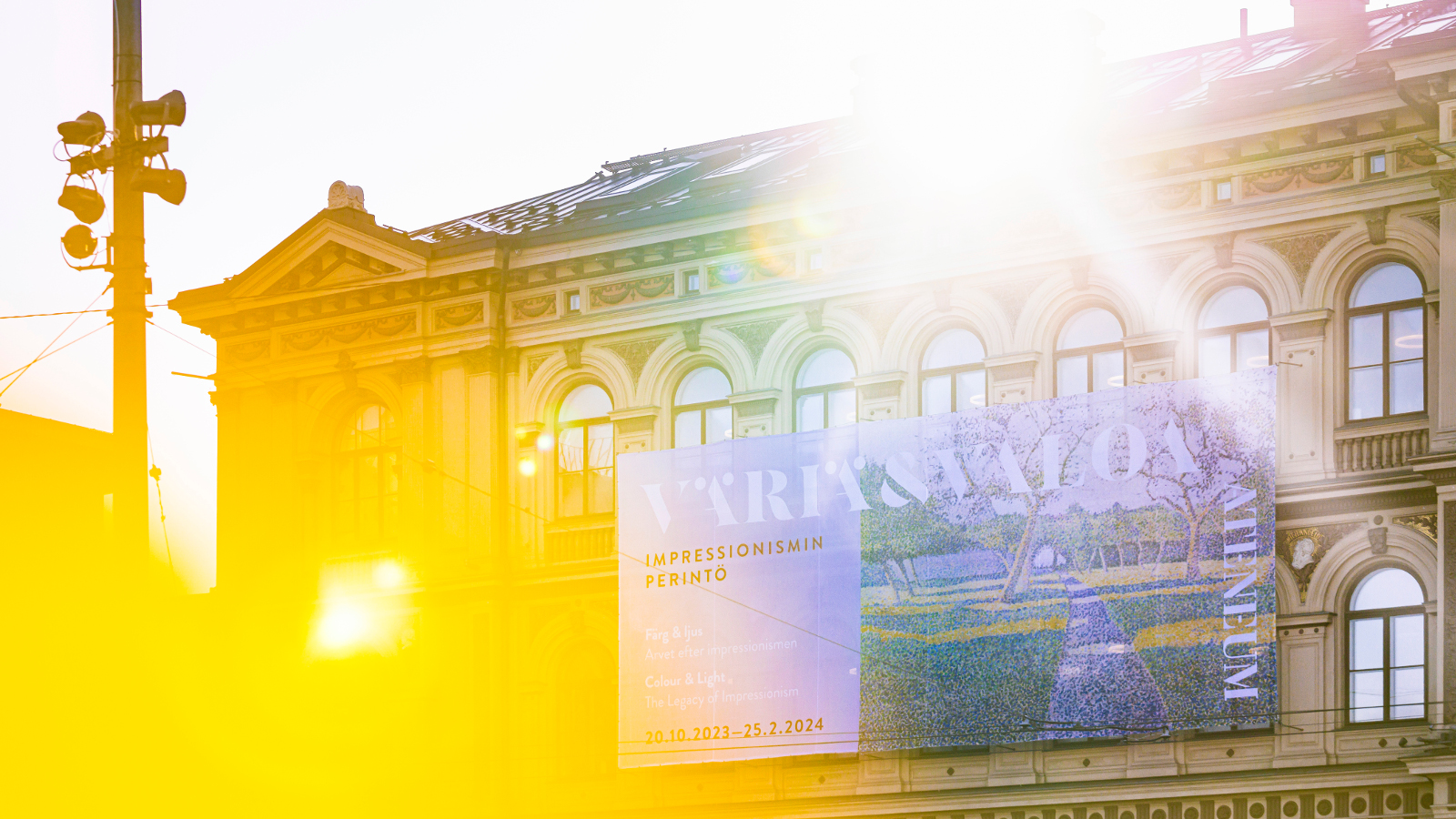 Kuvassa on Ateneumin päärakennuksen julkisivu ja siihen osuu keltainen valo vasemmalta ja oikealta tulee auringon valoa.  Seinällä on iso mainoslakana.