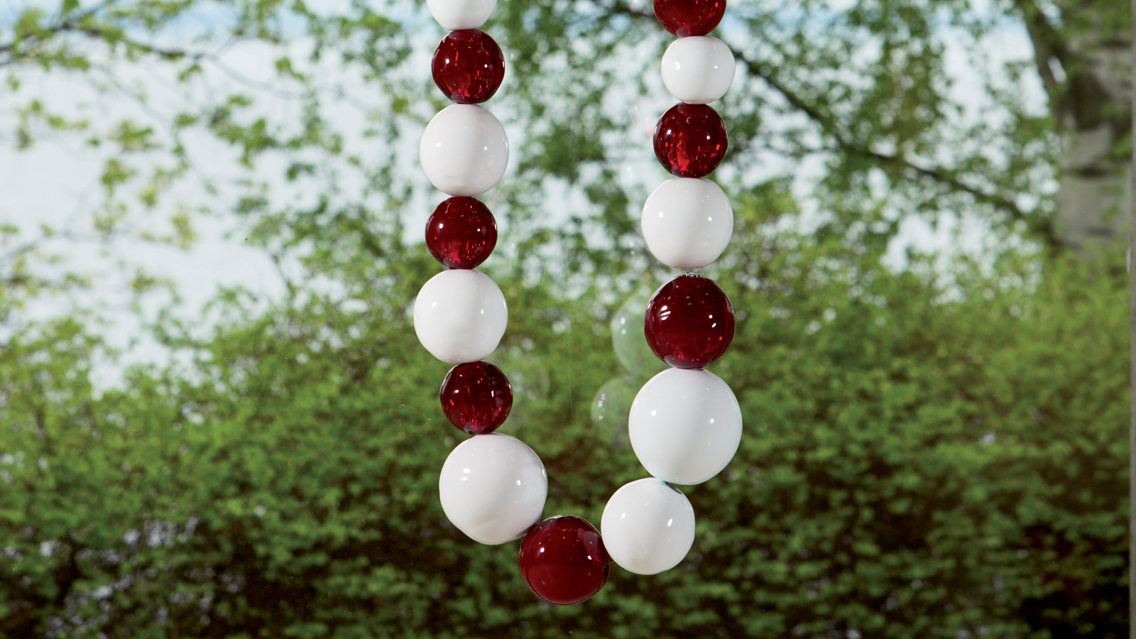 Kuvassa on isoja, valkoisia ja punaisia helmiä helminauhassa ripustettuna puun oksalle. Kuvassa näkyy helminauhan kaartuva pää.
