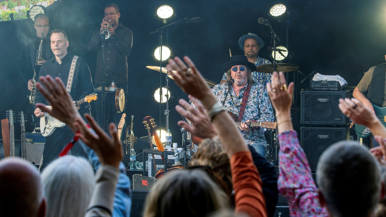 Kuvassa on Hector esiintymässä Salmelan lavalla ja taustalla on bändiä, jossa on kitaristi ja puhaltajia soittamassa.  Edessä on yleisöä kädet pystyssä ja näkyy päälakia. 