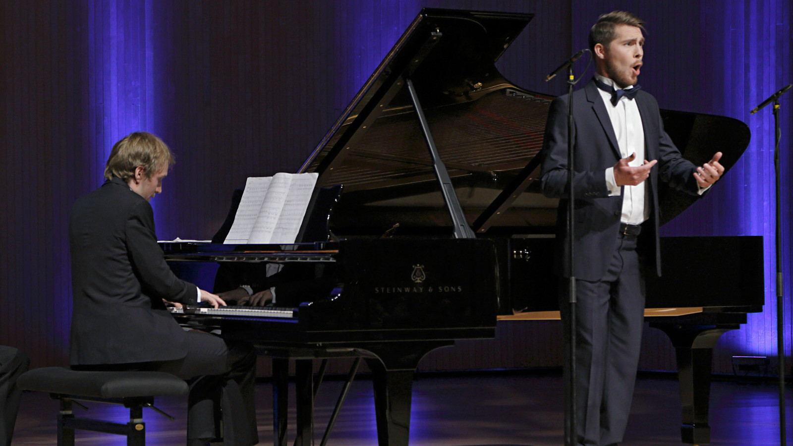 Kuvassa on sivuttain pianisti Juho Alakärppä flyygelin ääressä soittamassa.  Lavalla oikealla on laulamassa Aarne Pelkonen. Taustalla on sini-mustaa verhoa.