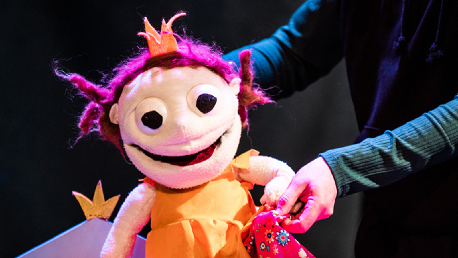 Kuvassa on Molla-Maijan näköinen nukke, jolla on oranssi paita ja punainen pystykö tukka.  Takaa näkyvät kädet ja tummaa taustaa.