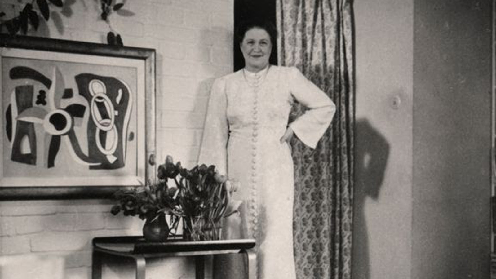 Kuvassa on Aino Aalto seisomamassa valkoisessa pitkässä, kangasnapitetussa puvussa. Vieressä on valkoista tiiliseinää ja siinä on abstrakti maalaus ja sen alla matala pöytä siinä on kaksi tulppaaniasetelmaa. Kuva on musta-valkoinen.