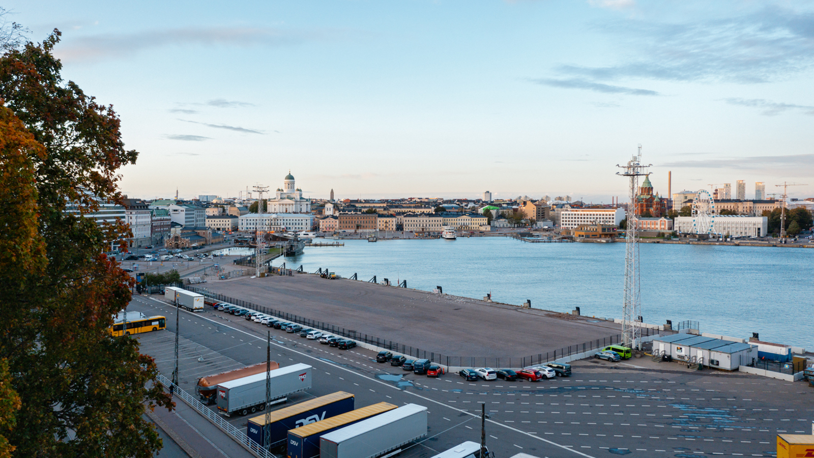 Kuvassa on Helsingin eteläsataman aluetta ja siinä näkyy tontti, jolle arkkitehtuuri- ja designmuseo tulee. Kuvassa näkyy kaukana tuomiokirkko ja kauppatoria sekä etualalla rekkoja parkissa. Taivas on sininen.