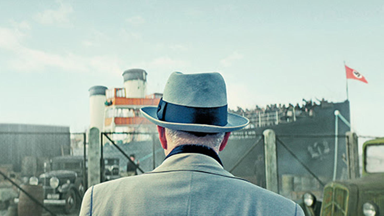 Kuvassa on mies takaapäin siten, että näkyy harmaata takkia ja hattu. Edessä näkuu satamassa olevaa laivaa ja sinistä taivasta.