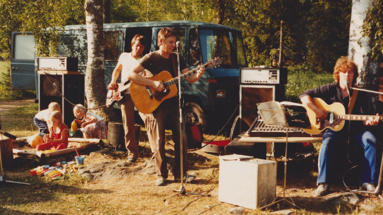 Kuvassa ovat bändin neljä jäsentä harjoittelemassa kesäisessä ympäristössä koivujen alla.  Heistä näkyy kolme ja heillä on kitarat  ja he soittavat. Vasemmalla on lapsia leikkimässä hiekkalaatikolla.