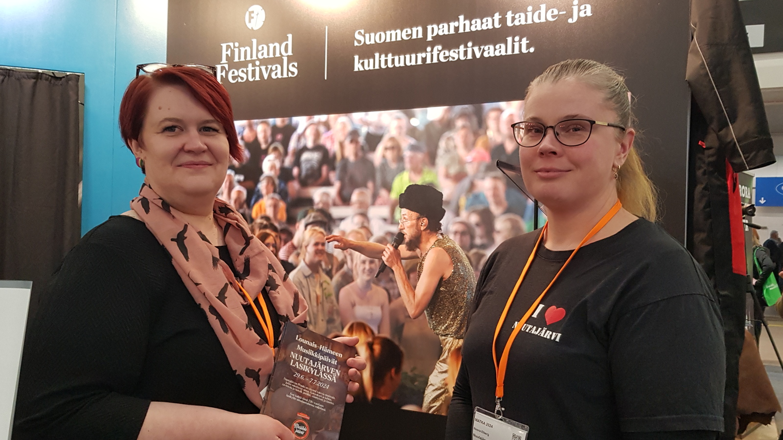Kuvassa ovat Päivi Lehti ja Noora Elberg mustissa t-paidoissa. Taustalla näkyy Finland Festivals -teksti ja festarikuva, jossa on paljon ihmisiä.