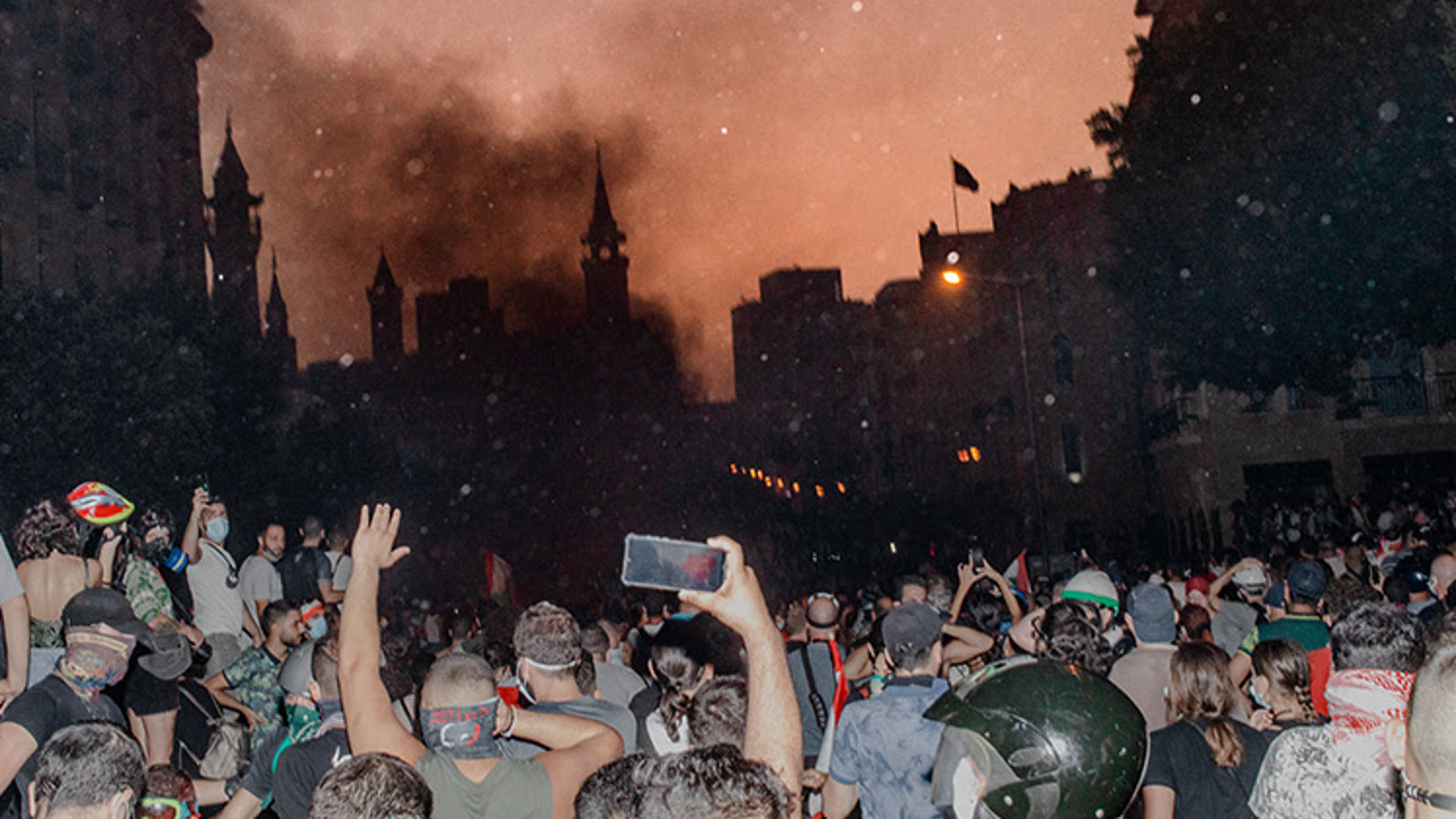 Kuvassa on alaosassa väkijoukkoa ja jotkut nostavat käsiää ja jotkut kuvaavat kännykällä edessä olevaa savuavaa kaupunkia. Sieltä nousee ruskeata savua.