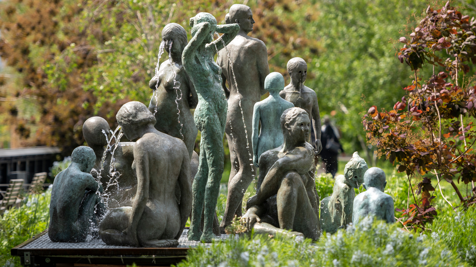 Kuvassa on ryhmä patsaita naisista ja lapsista selin seisomassa ja istumassa vihreiden lehtipuiden ja pensaiden keskellä suihkulähteen äärellä. Patsaat ovat vihreän harmaita.