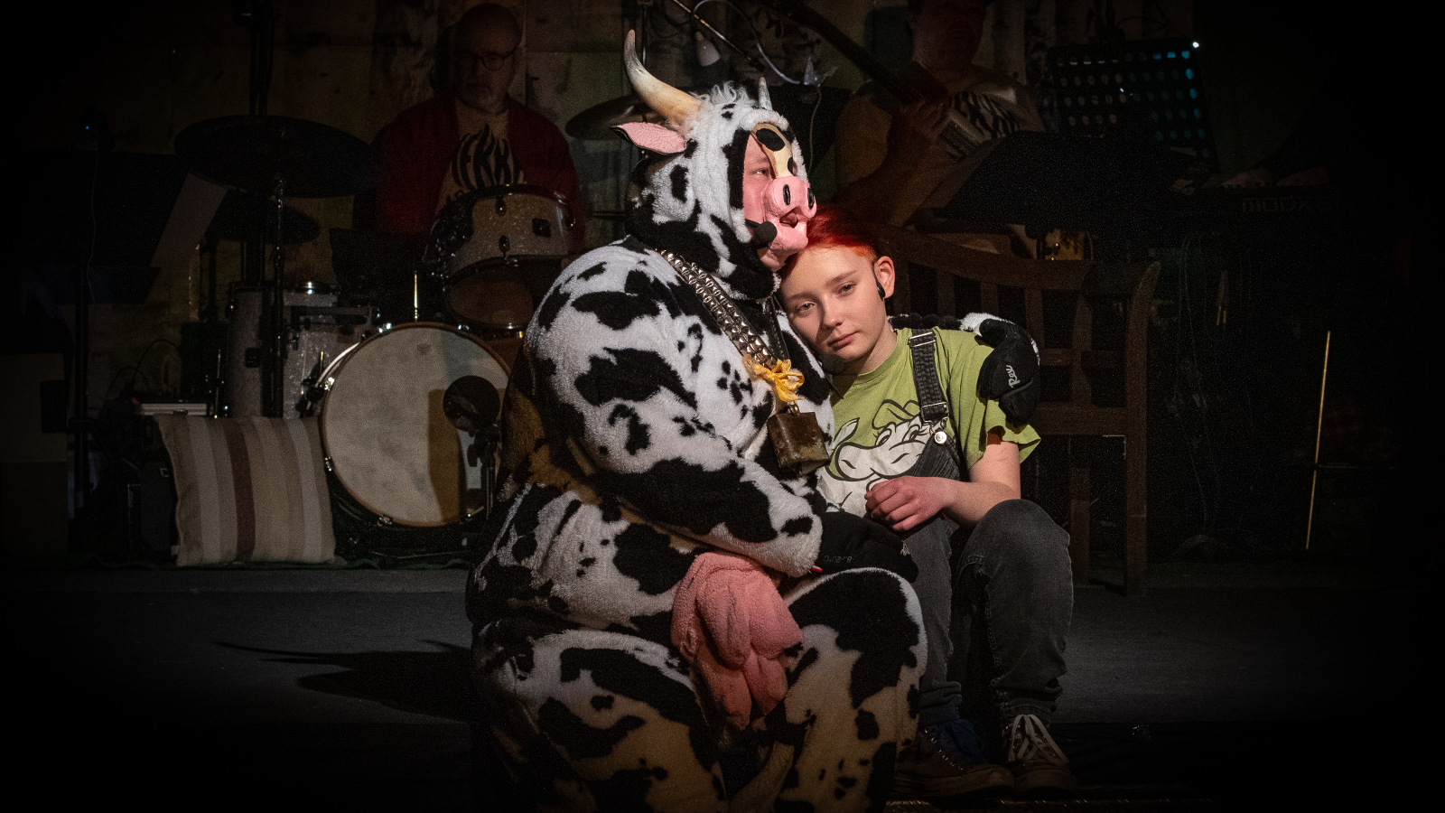 Kuvassa on istumassa musta-valkoisessa lehmäasussa oleva näyttelijä ja häneen nojaa nuori tyttö vihreässä paidassa. Tausta on tumm ja valaistus kohdestuu heihin.