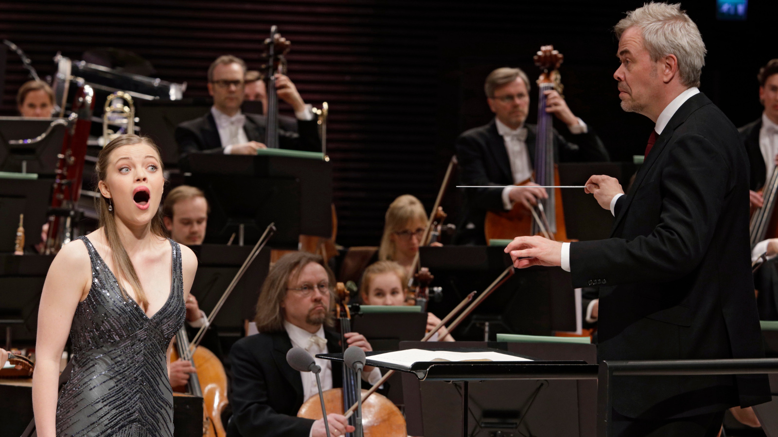 Kuvassa on laulamassa Johanna Wallroth vasemmalla mustassa, kiiltävässä hihattomassa puvussa ja taustalla on orkesteri.  Oikealla selin on Hannu Lintu johtamassa orkesteria.
