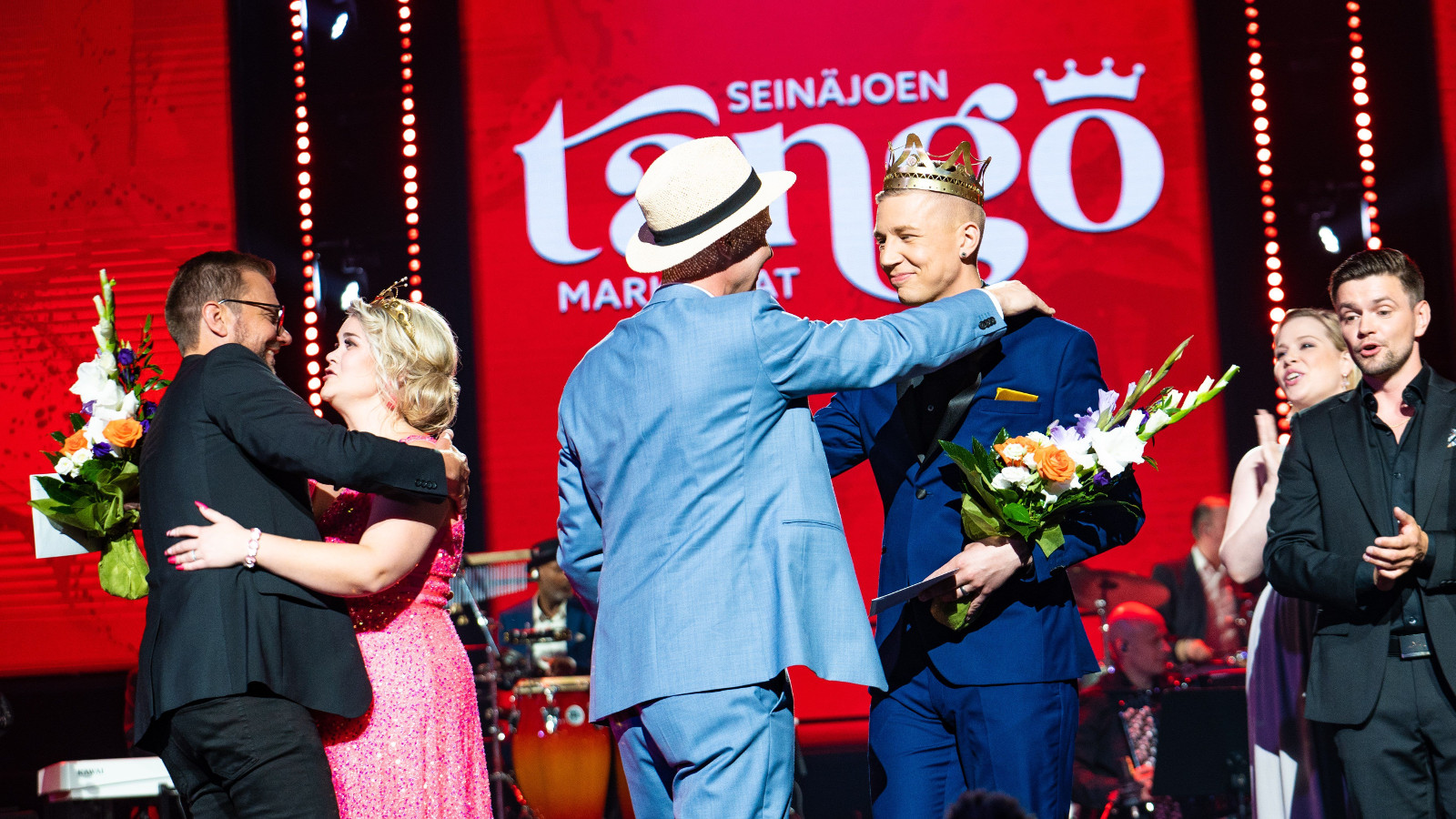 Kuvassa on menossa Tangomarkkinoiden voittajien onnittelut ja kukitus. Vasemmalla on Charlotta Saaren onnittelu ja halaus.  Hänellä on pinkki puku päällään.  Vieressä on Pasi Flodströmin onnittelut ja onnittelijalla on vaalean sininen puku.  Tausta on punainen ja seinällä lukee tango.