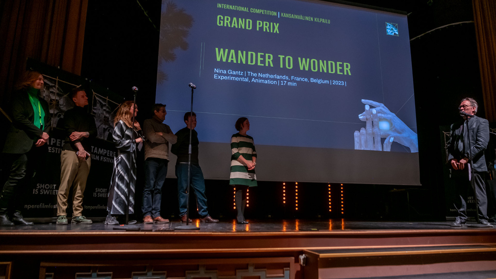 Kuvassa on rivissä seisomassa näyttämöllä kuusi palkinnonsaajaa ja oikealla on palkintojen jakaja. Taustalla on iso näyttö, jossa lukee isoin kirjaimin Grand Prix ja Wander to Wonder. Tausta on tumman sininen.