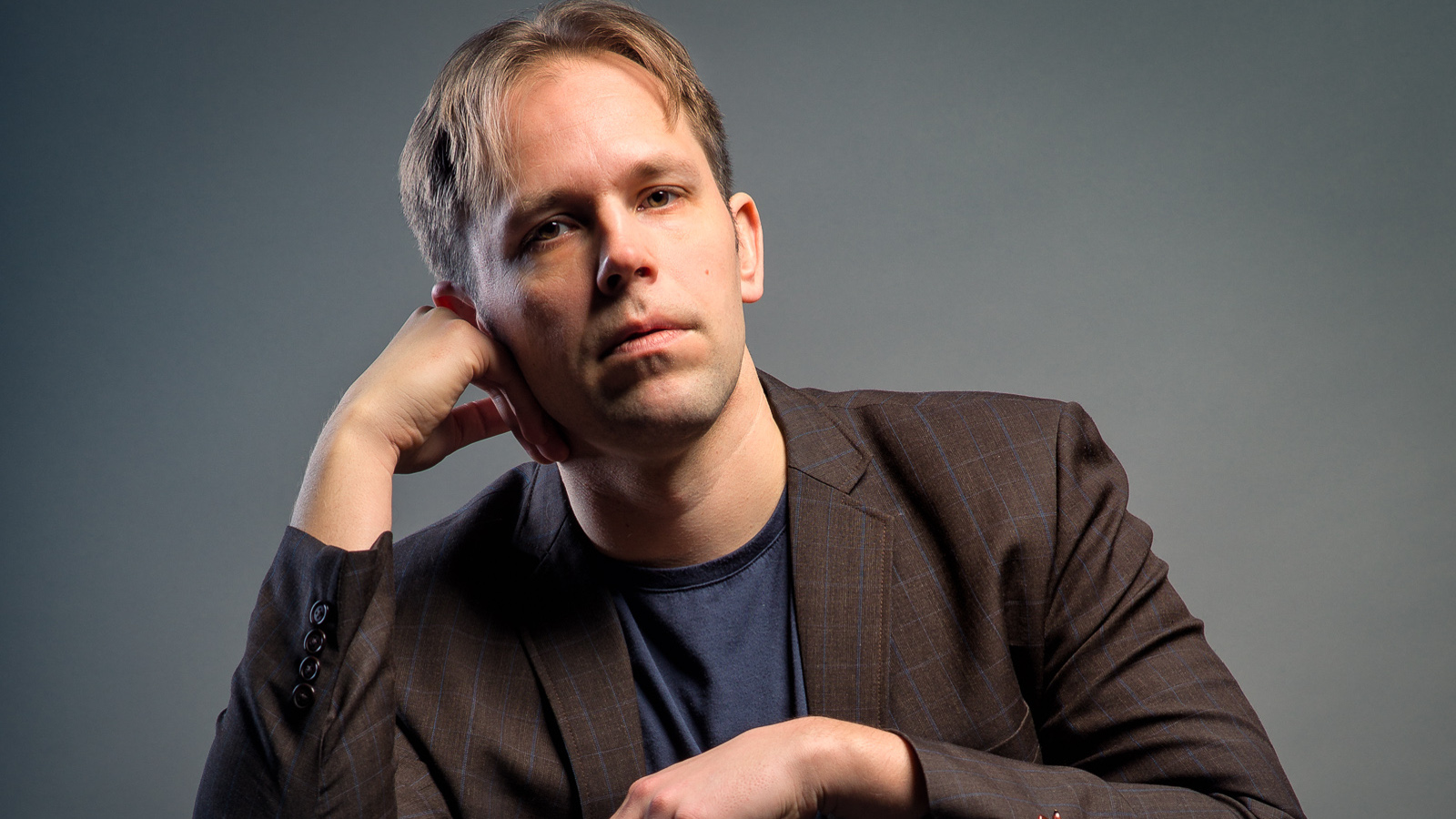 Kuvassa on Jukka Merjanen ruskehtavassa takissa ja hän nojaa oikeaan käteensä. Tausta on harmaa.