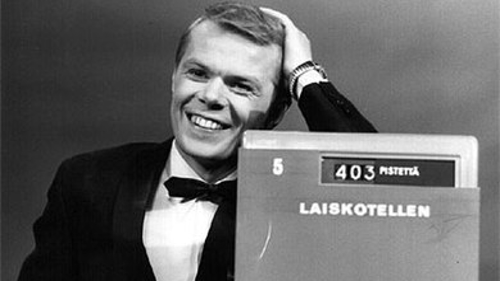 Kuvassa on Lasse Mårtenson mustassa takissa ja rusetissa puolivartalokuvassa. Han nojaa vasemmalla kädellään kassakoneeseen ja siinä näkyy teksti Laiskotellen ja 403 pistettä sekä numero 5.