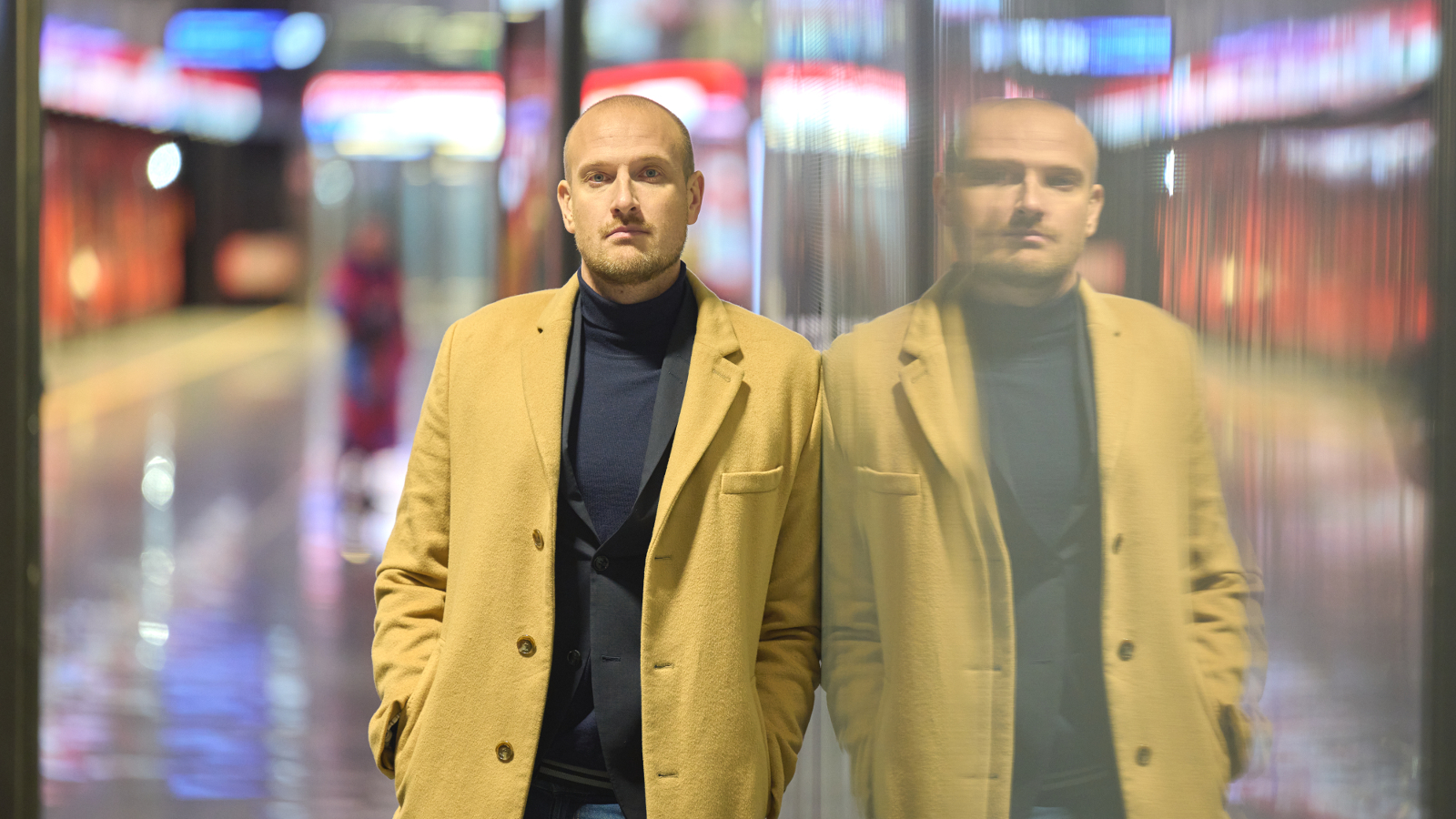 Kuvassa on Max Seeck keltaisessa takissa ja taustalla on himmennettynä Helsingin metroasemalaituria. Seeck on kahdennettu oikealle himmeämmin kuin vasemmalla eli on peilikuvassa.