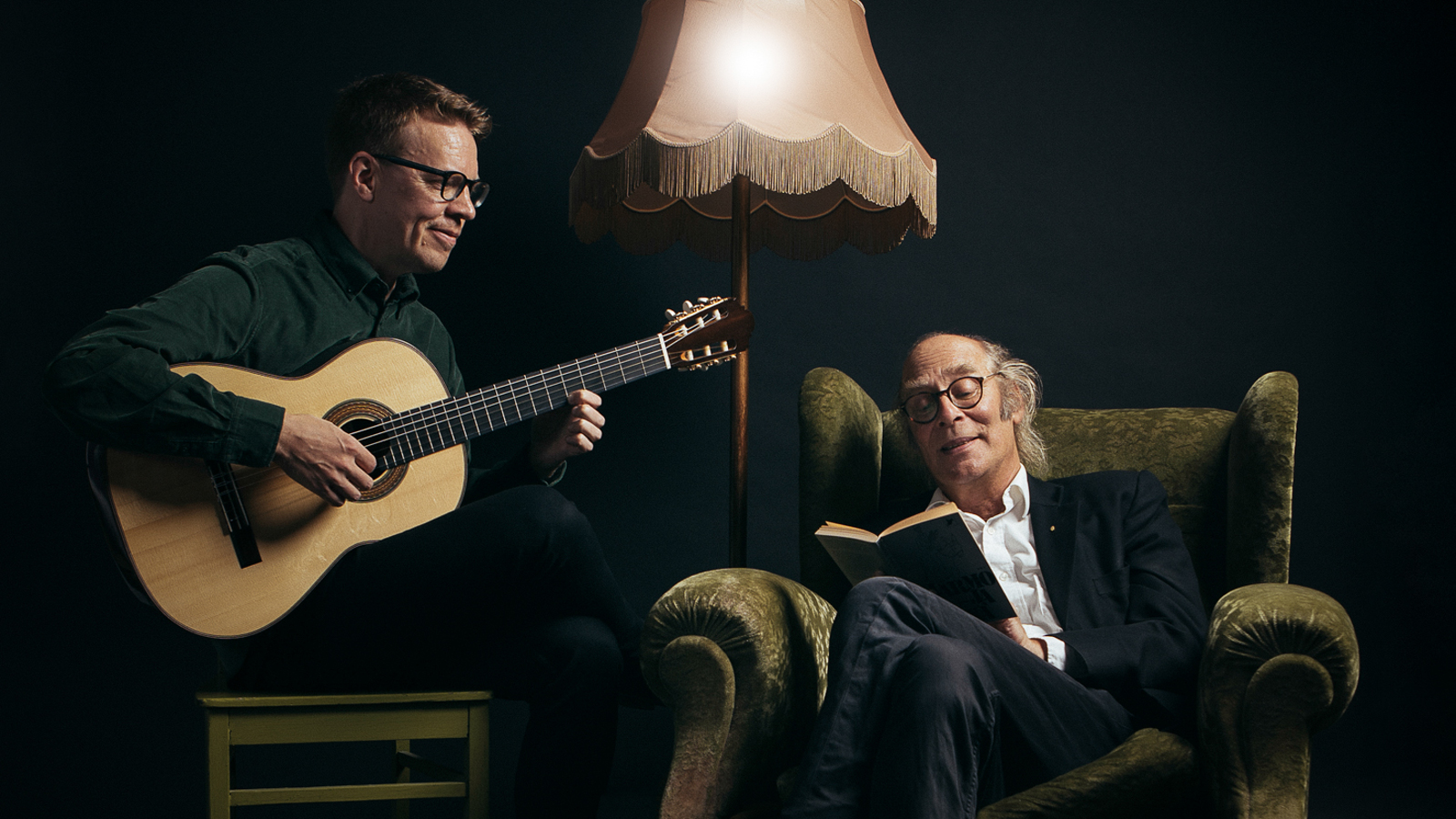 Kuvassa on vasemmalla Petri Kumela soittamassa kitaraa ja oikealla Vesa Vierikko istumassa isossa nojatuolissa. Taustalla on tummaa huonetta.