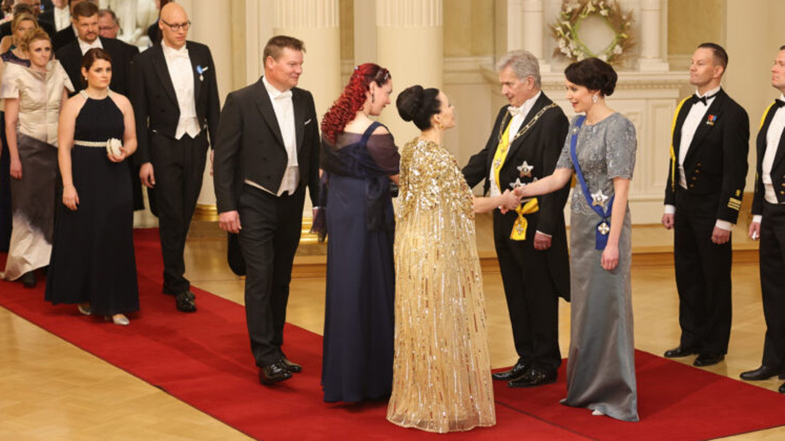 Kuvassa ovat oikealla Jenni Haukio ja Sauli Niinistö vastaanottamassa vieraita ja kättelemässä on melkein selin nainen punaisissa, kiharaisisssa hiuksissa ja mustassa pitkässä mekossa.  Hänen takanaan on mies mustassa frakissa.  Hänen takanaa näkyy pariskuntia tulossa kättelyyn punaisella matolla.  Lattia on ruskehtavaa parkettia. Presienttiparin takana on kaksi adjutanttia mustissa kadettiasuissa.