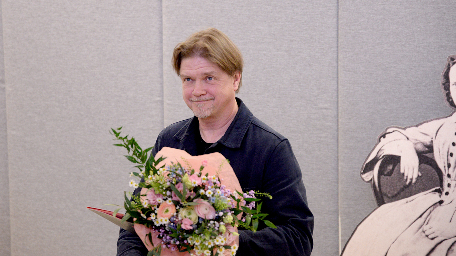 Kuvassa on Timo Parvela vaaleanpunainen kukkakimppu kädessään ja mustassa takissa.  Tausta on harmaa seinä.  Oikealla näkyy osittain isoa Minna Canthin piirroskuvaa.