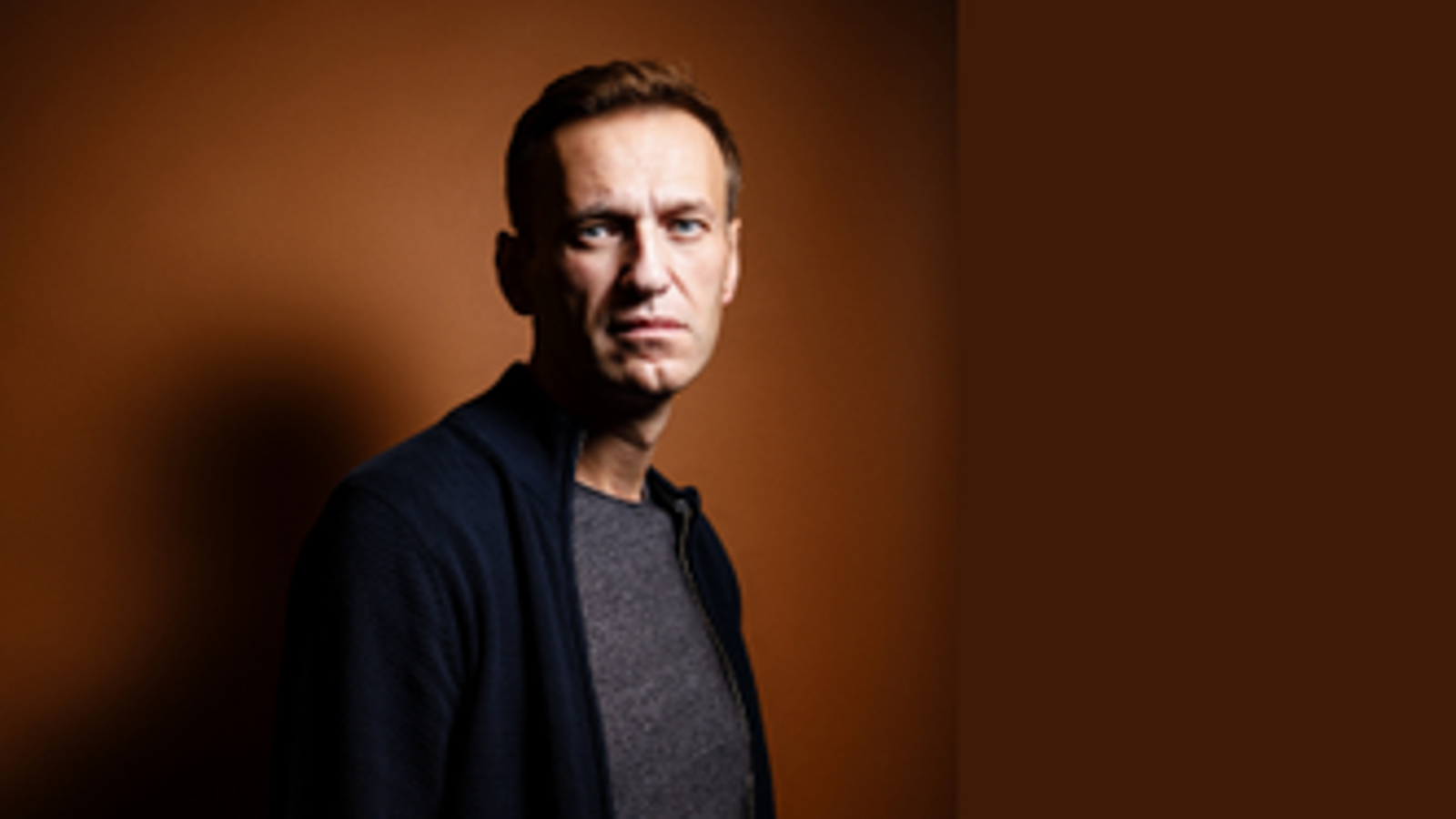 Kuvassa on Aleksei Navalny puolivartalokuvassa seisomassahieman vinottain. Hänellä on musta villatakki ja harmaa paita päällään. Tausta on ruskea.