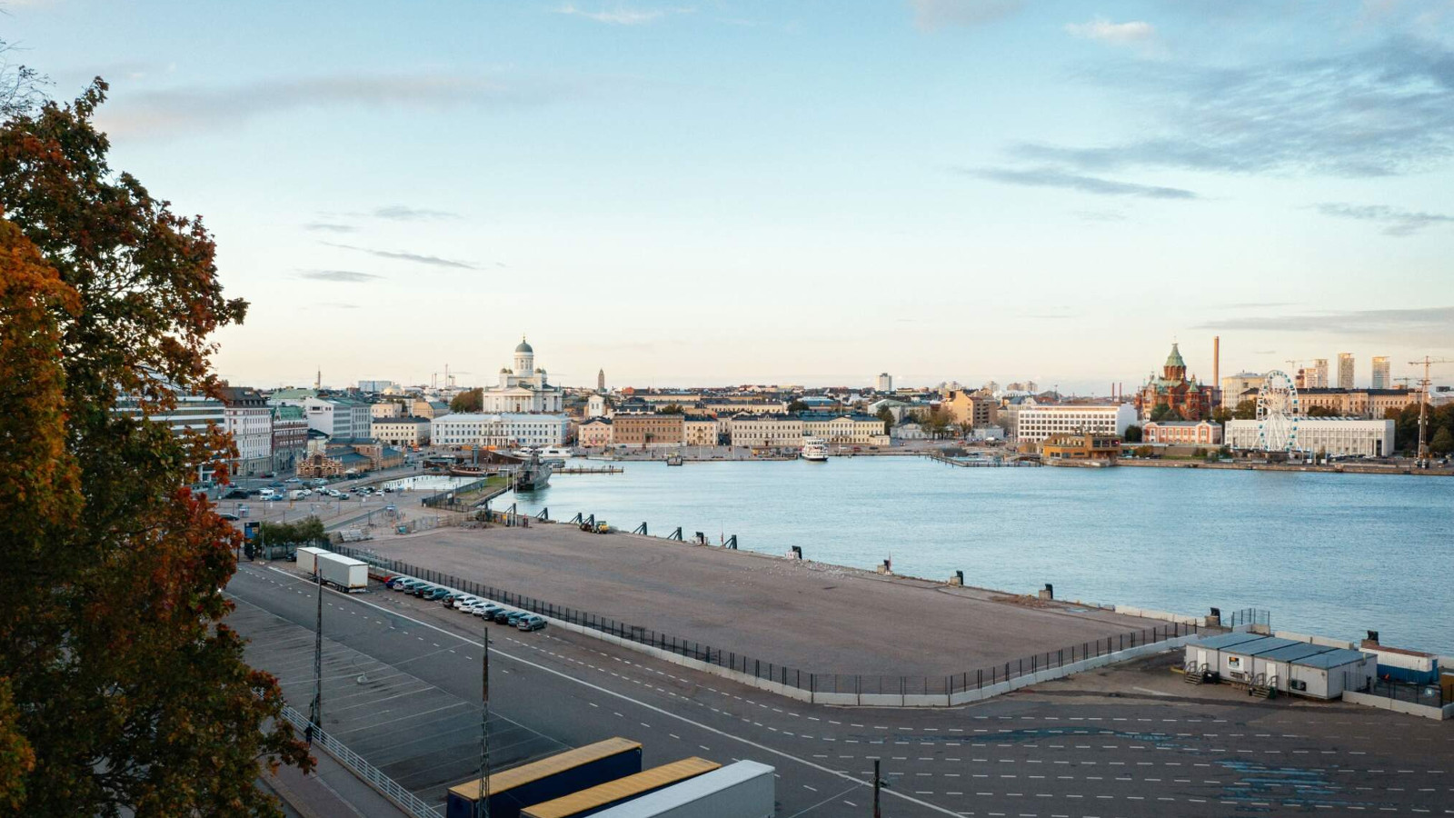 Kuvassa on taaempana Helsingin kauppatorin rakennukset ja edustalla näkyy sinistä merenlahtea.  Edessä on asfalttikenttää ja ylhäältä tulee leveät A-kirjaimen muodot tekevät palkit.