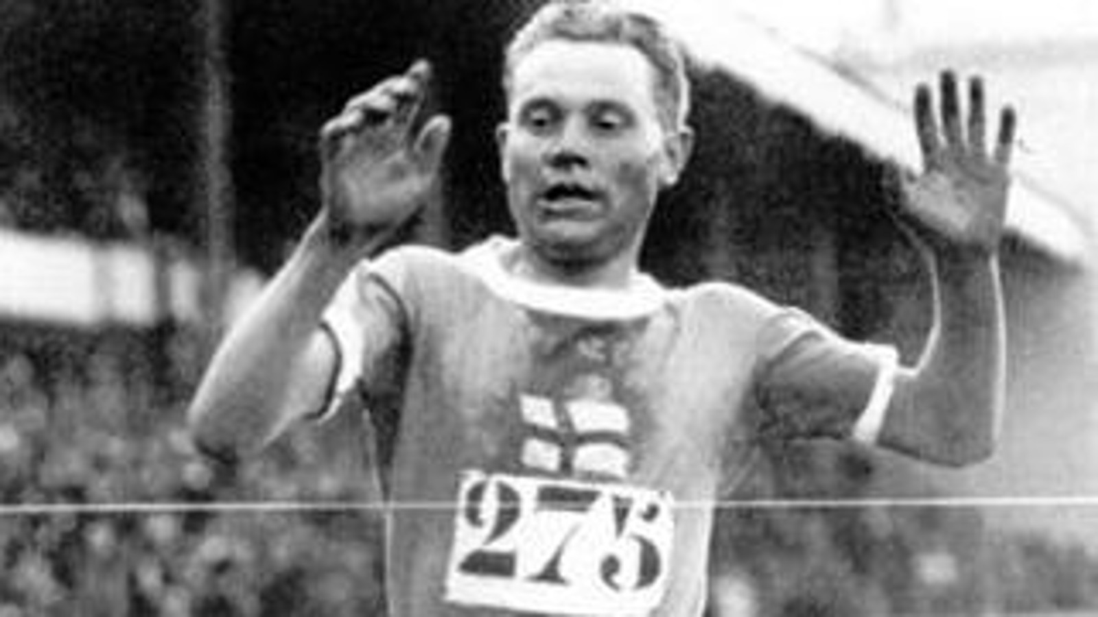 Kuvassa on Paavo Nurmi tulossa maaliin.  Hänellä on rinnassaan on numero 275. Hänen edessään on maalinauha.  Taustalla on himmeästi yleisöä.  Nurmella on kädet kohotettuna ylös.