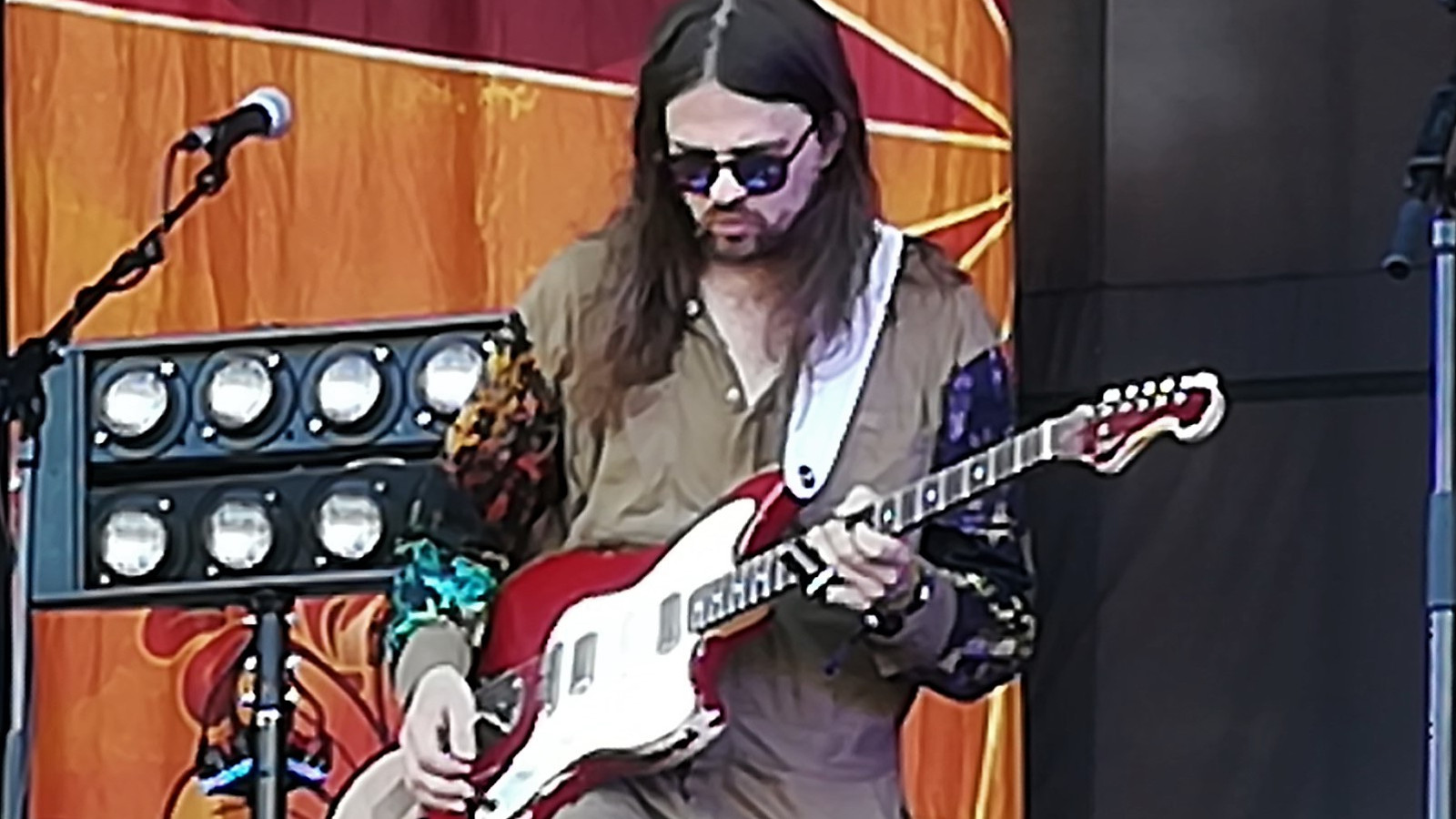 Kuvassa on Timo Kämäräinen soittamassa kitaraa ja hän katsoo alaspäin. Hänellä on pitkät tummat hiukset ja harmaa paita päällään. Taustalla on oranssia seinää ja edessä on kahdeksan valon taulu. Oikealla on mustaa taustaa.
