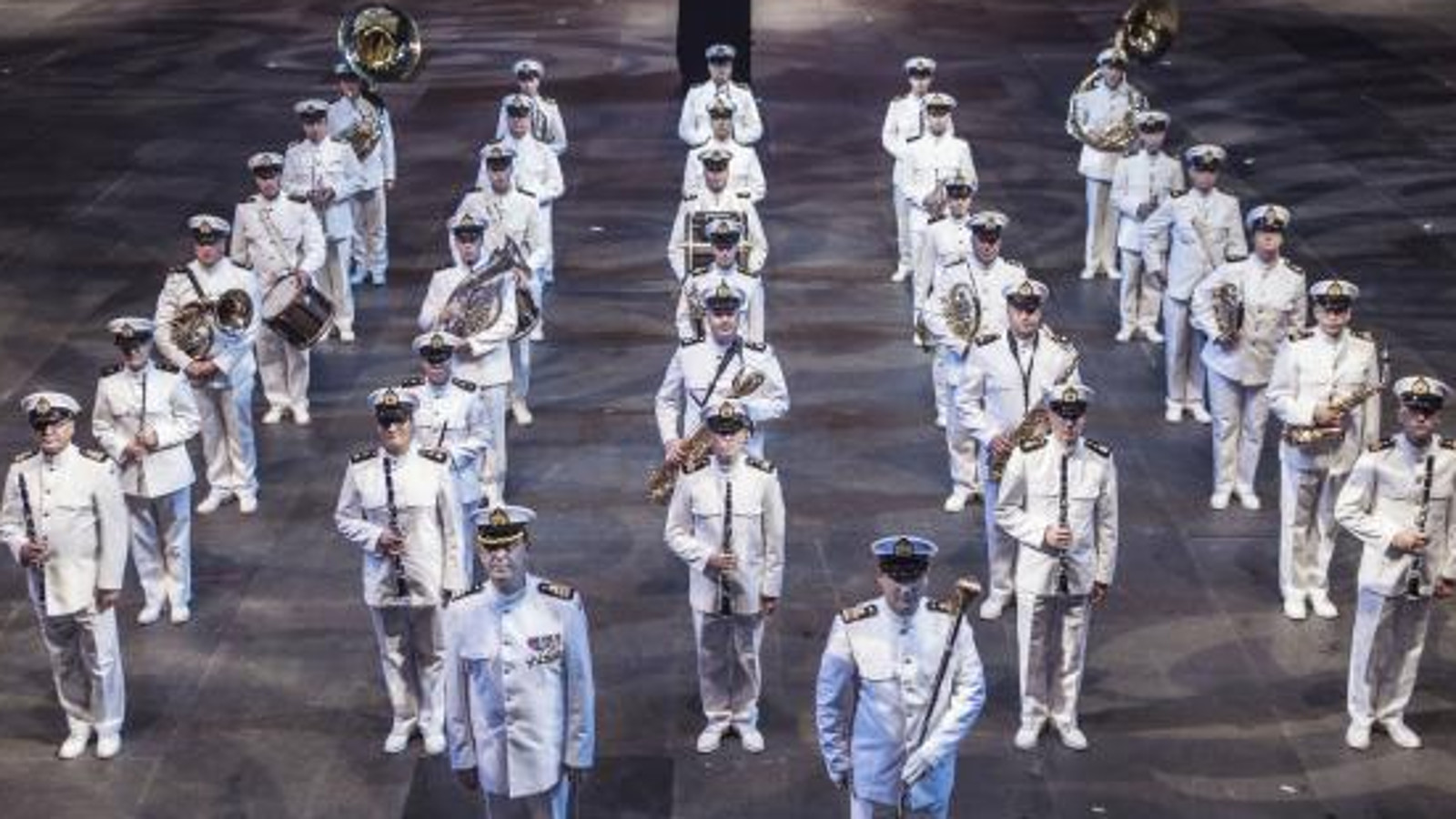 Kuvassa on Laivaston soittokunta valkoisissa puvuissa ja lakeissa seisomassa viidessä rivissä soittimet kädessään.  Kuva on otettu yläviistossa. Lattia on harmaa.