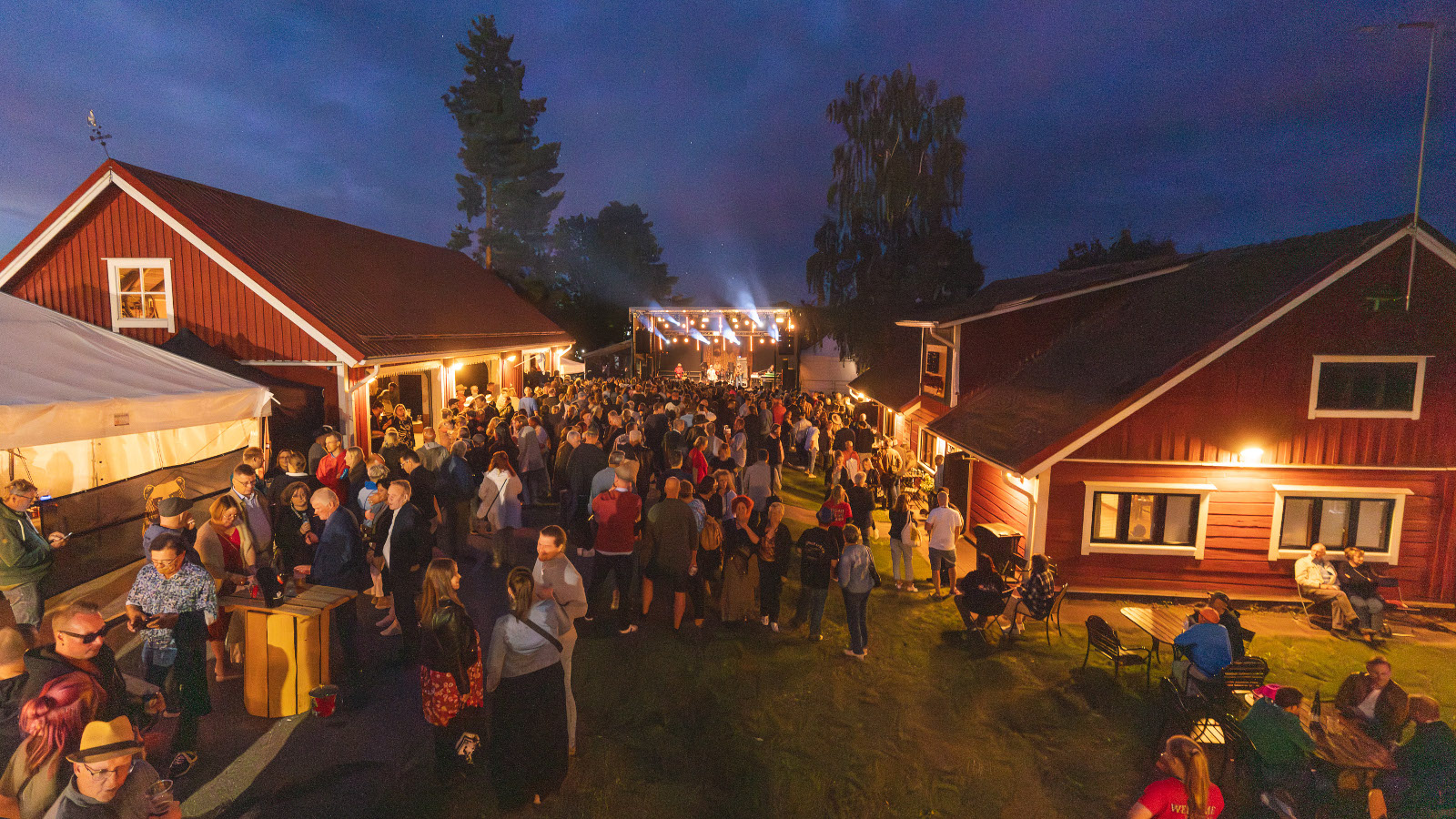 Kuvassa on maatalon pihapiiriä, jossa on punavalkoisia rakennuksia ja vasemmalla valkoinen teltta.  Pihapiirissä on paljon ihmisiä rakennusten välissä seisomassa ja taustalla on tumman sinistä iltataivasta loppukesällä.
