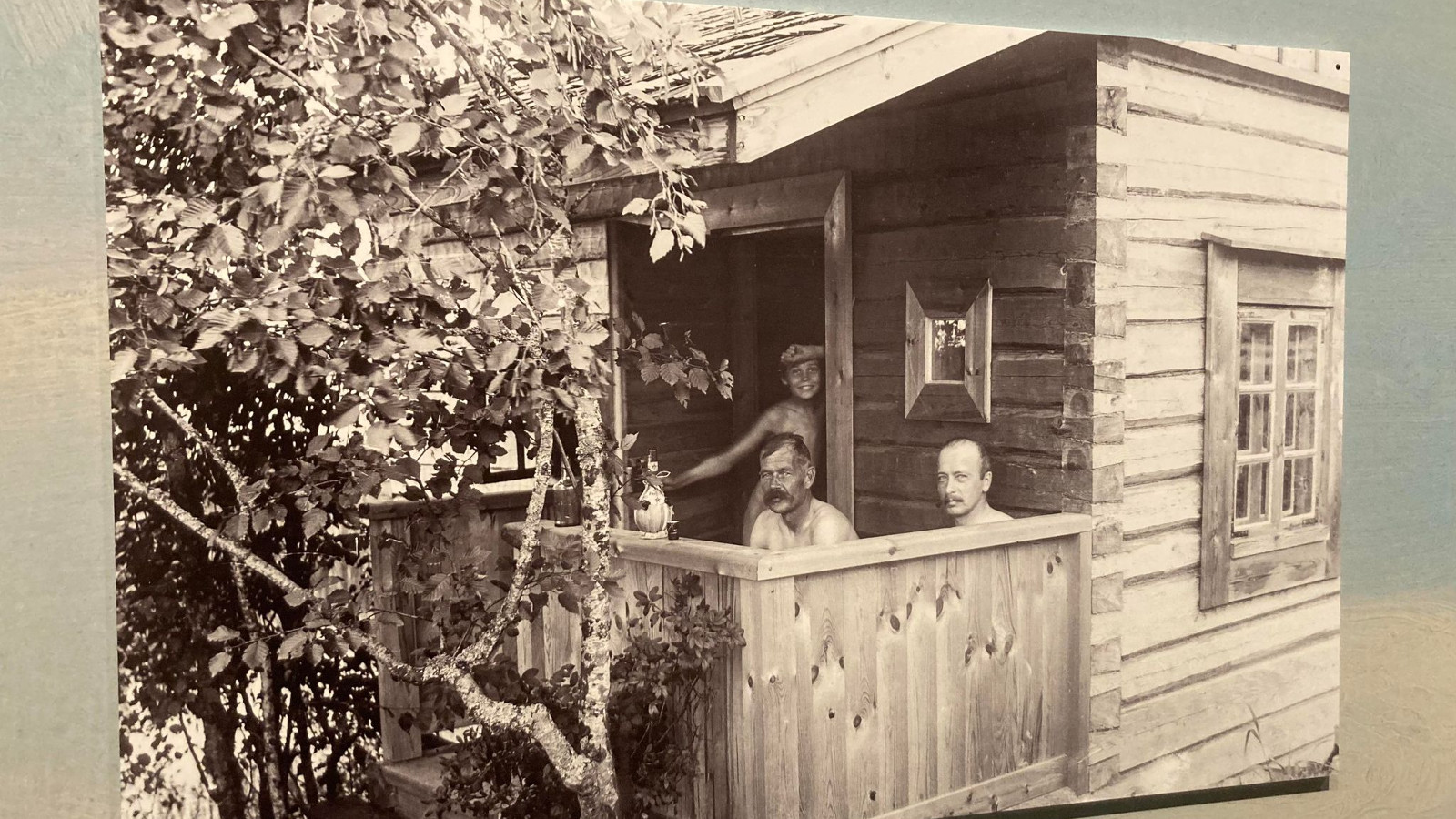 Kuvassa on hirsisauna ja sen edessä aidattu terassi. Sieltä näkyy kahden miehen päät ja sisältä ovalta katsoo nuori poika. 